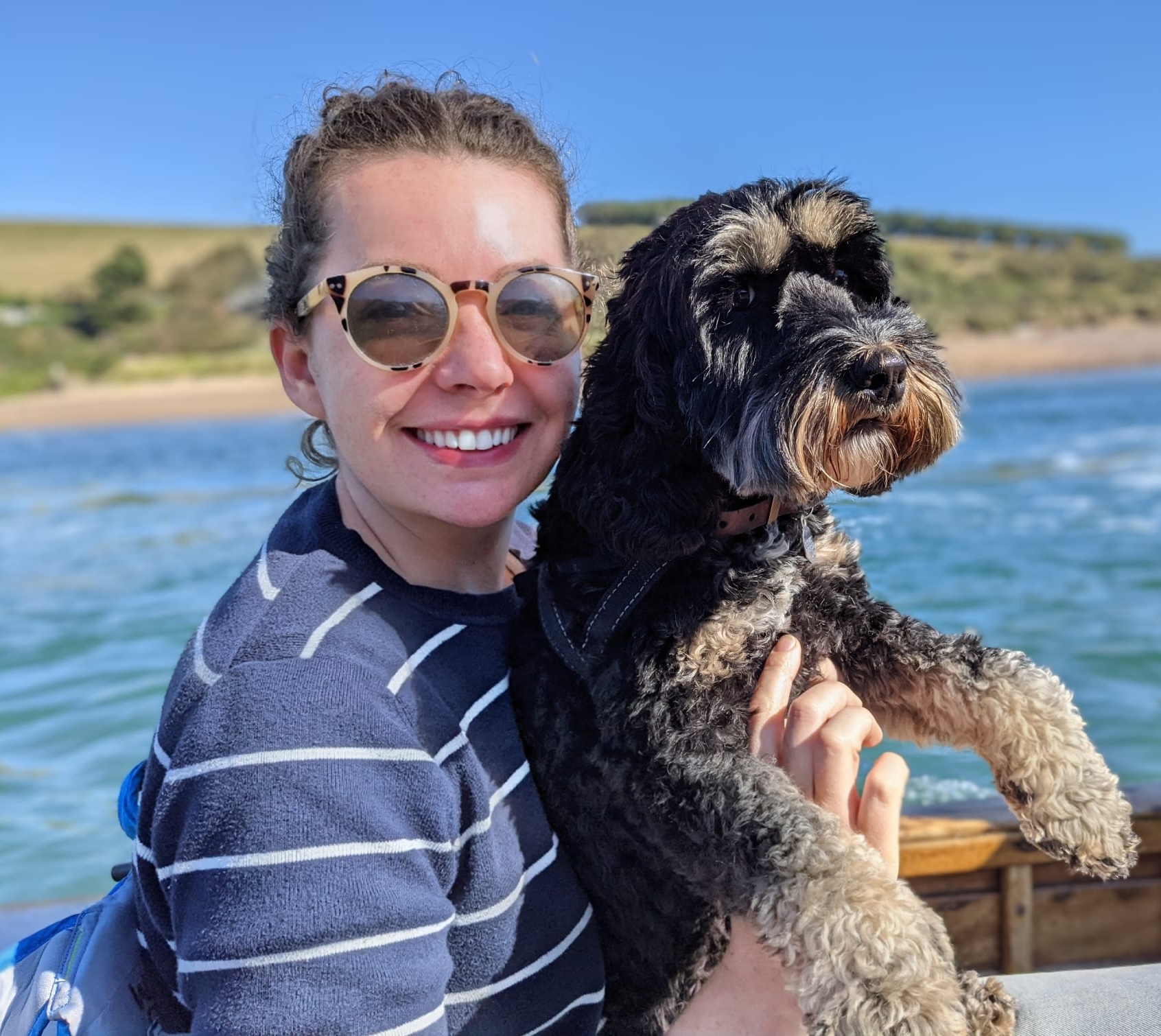 Smiling woman wearing round sunglasses and a navy blue shirt with white stripes holds a black and beige curly-haired dog while sitting on a boat with water and a grassy shore in the background under a clear blue sky.