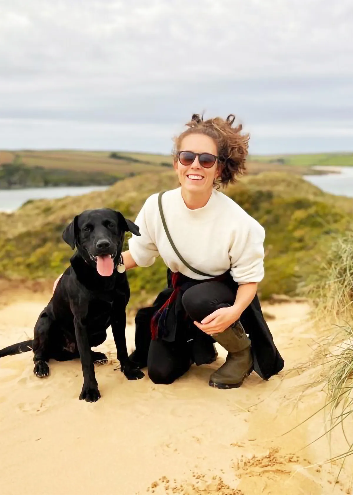 Woman wearing sunglasses, a white sweater, black pants, and boots kneeling on sandy ground next to a black dog with its tongue out, with grassy hills and water in the background.