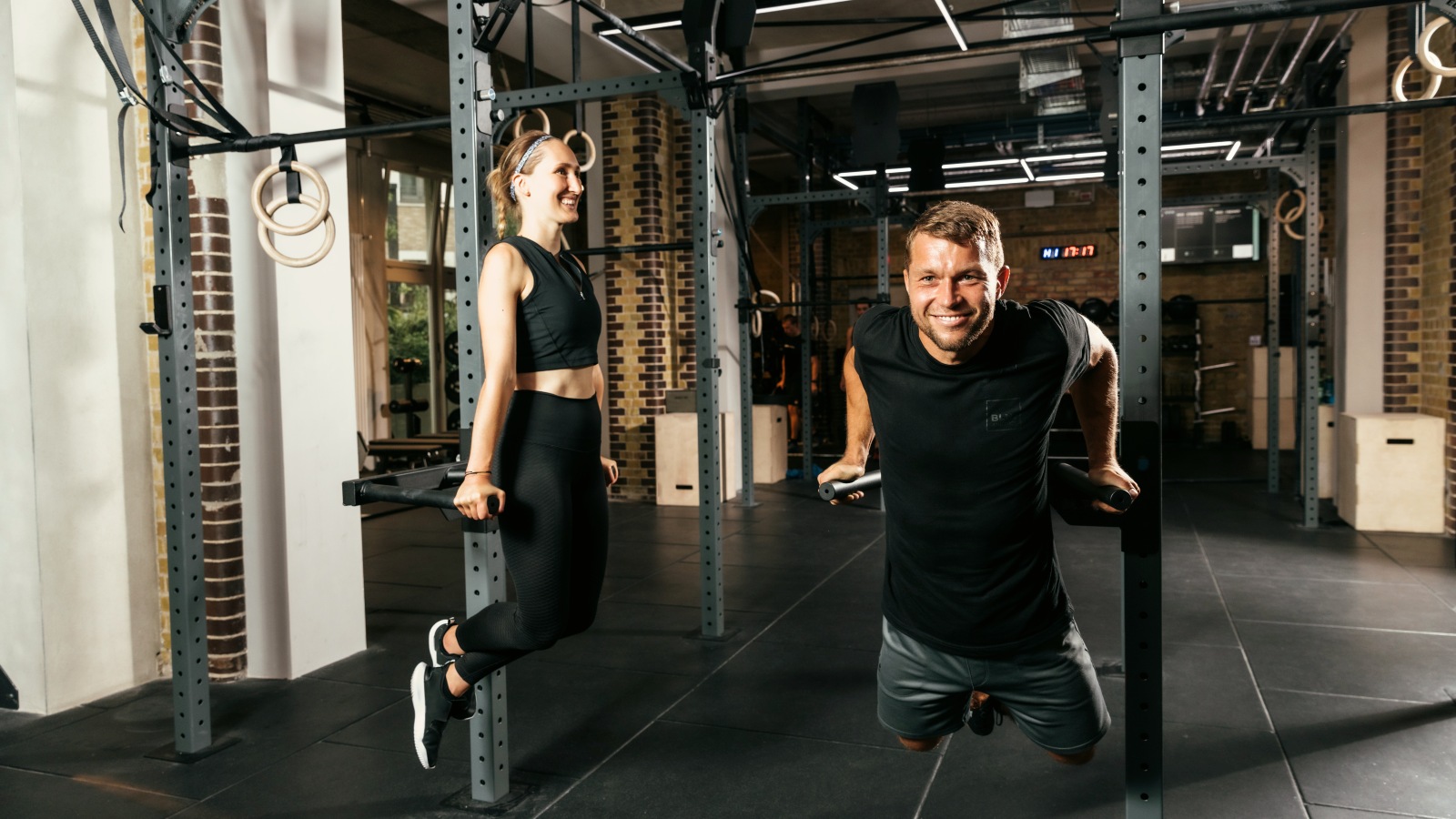 Eine Frau und ein Mann machen lachend Dips an den Griffen des Rigs bei BLN.Athletics, über ihnen hängen Turnringe.