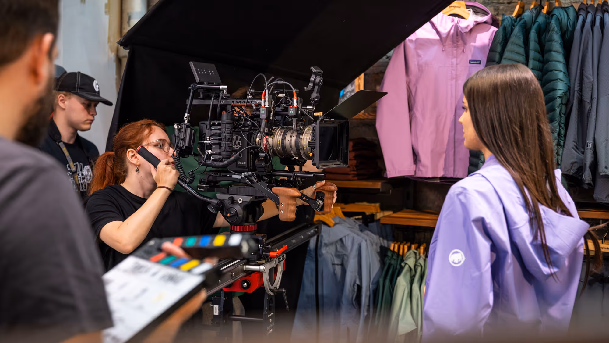 Film crew filming a woman in a lavender jacket inside a clothing store with outdoor jackets displayed.