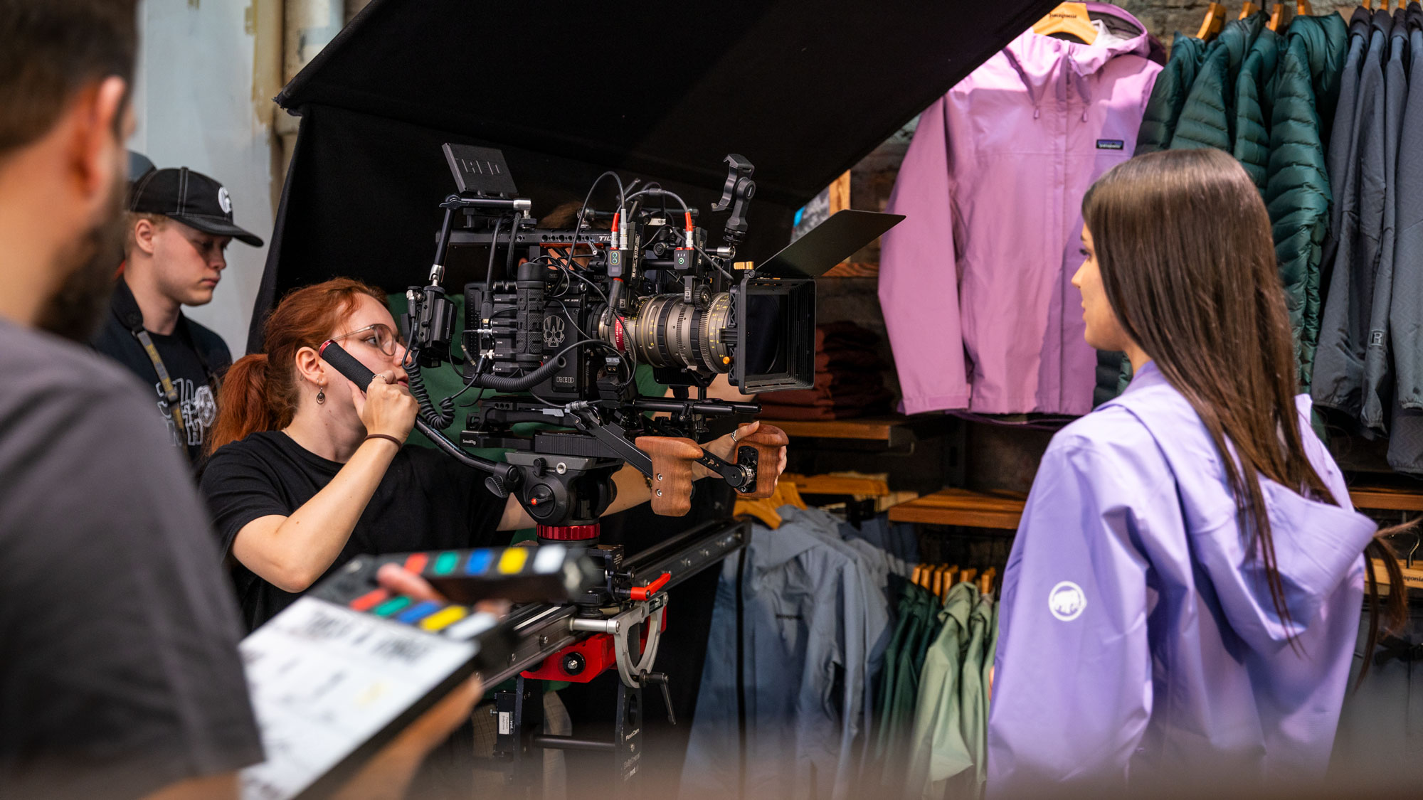 Film crew filming a woman in a lavender jacket inside a clothing store with outdoor jackets displayed.