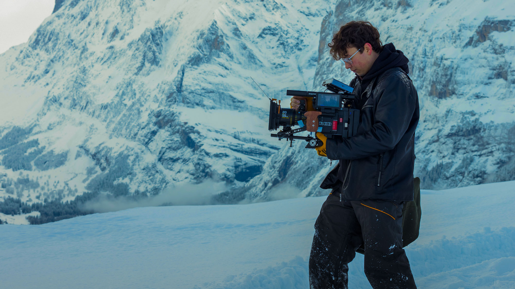 Person in winter clothing operating a professional video camera in a snowy mountain landscape.