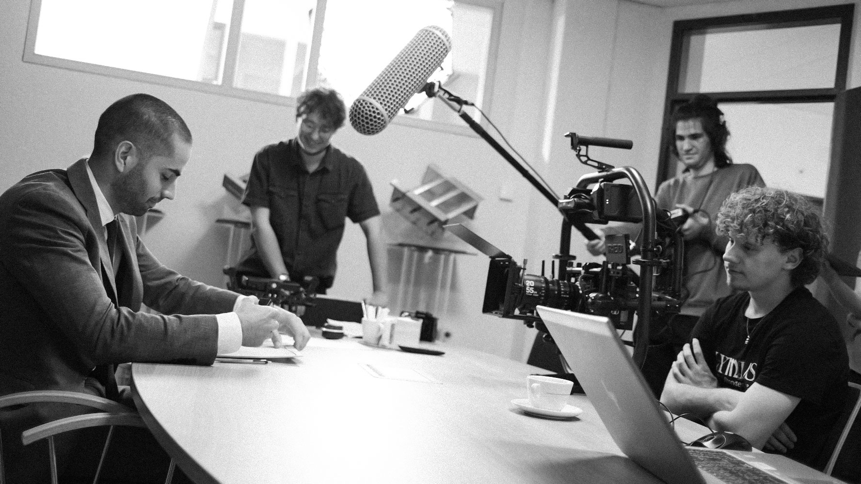 Black and white photo of a film crew recording a man in a suit at a table, with a boom microphone, camera, and laptop visible.