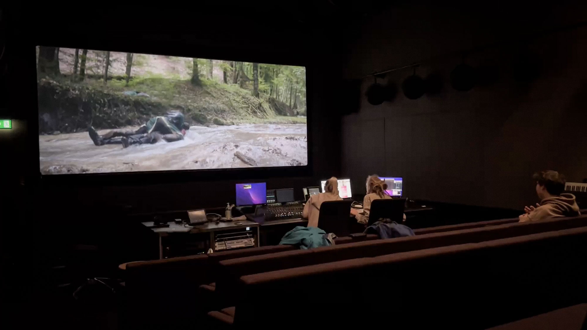 A dark post-production studio with three people seated, two working at computers and one watching a large screen showing a person lying in a river.