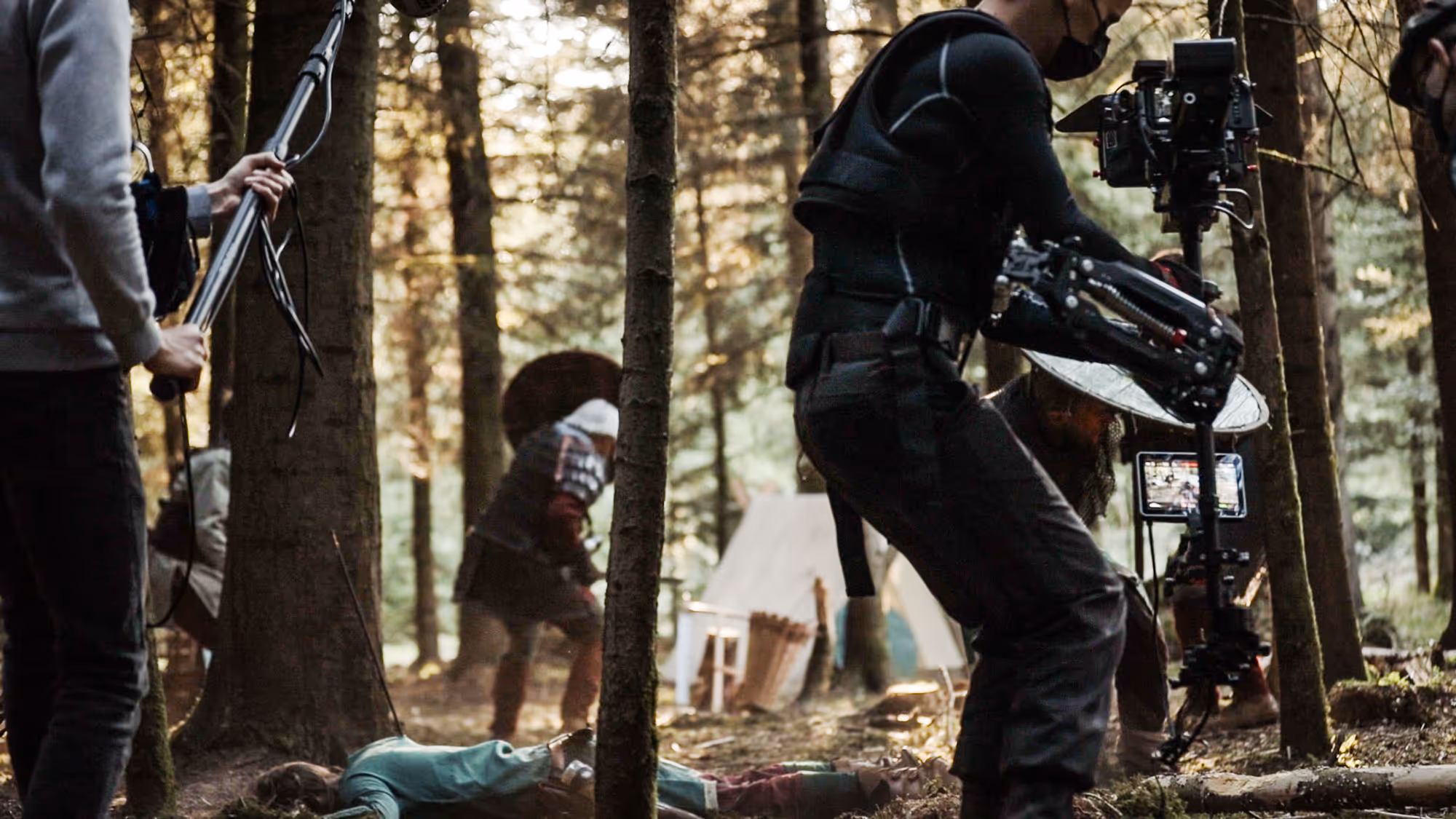 Film crew with camera and boom mic capturing a battle scene in a forest with actors in medieval armor.
