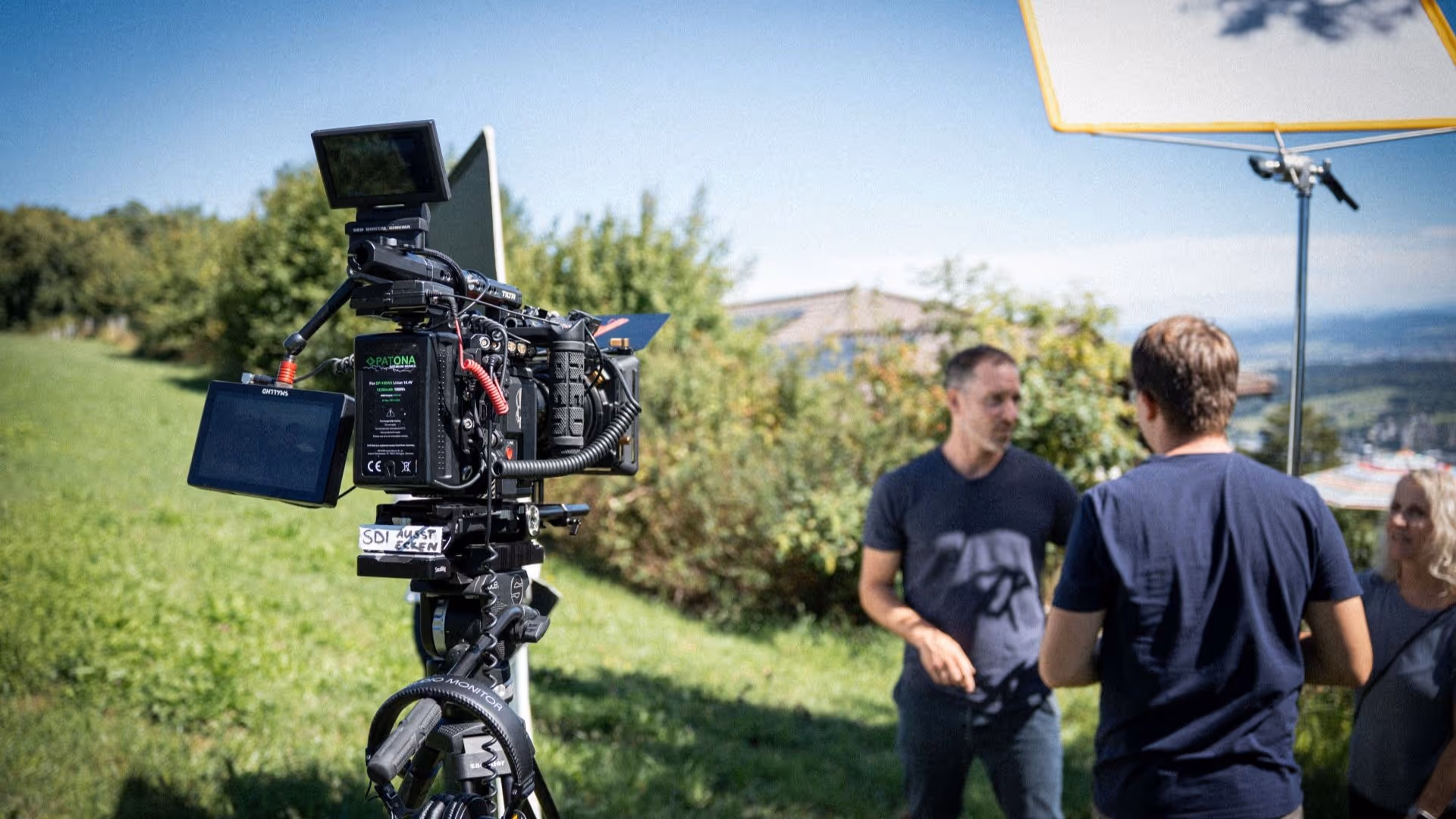 BTS Still einer Videoproduktion für B2B Marketing.
Links sieht man eine Filmkamera Red Dragon 6K mit Zubehör. Rechts unscharf imHintergrund 3 Personen welche miteinander sprechen. Über ihnen ist ein Difusor auf einem C-Stand zu sehen.