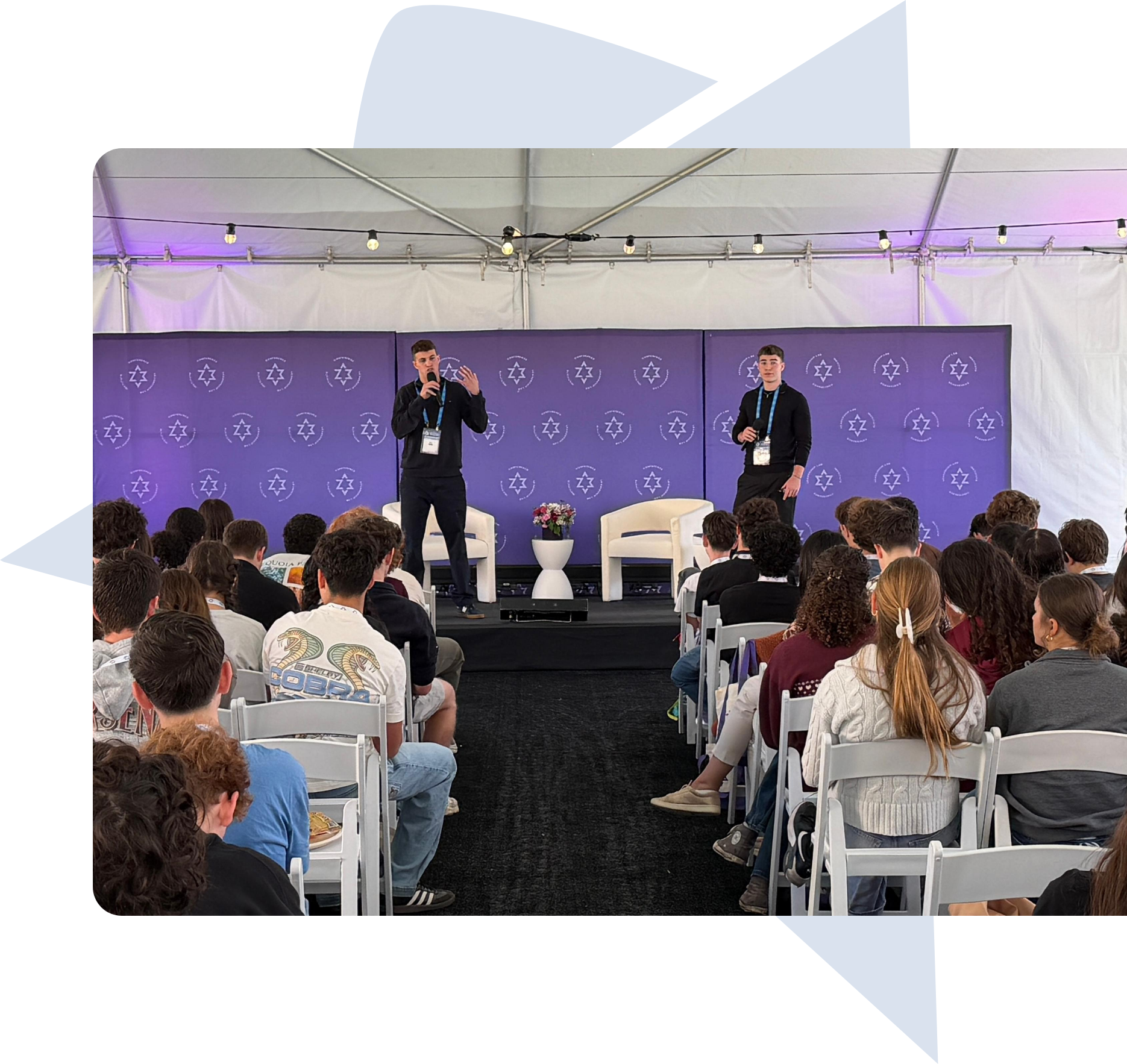 Two men standing on stage speaking to an audience seated in white chairs under a tent.