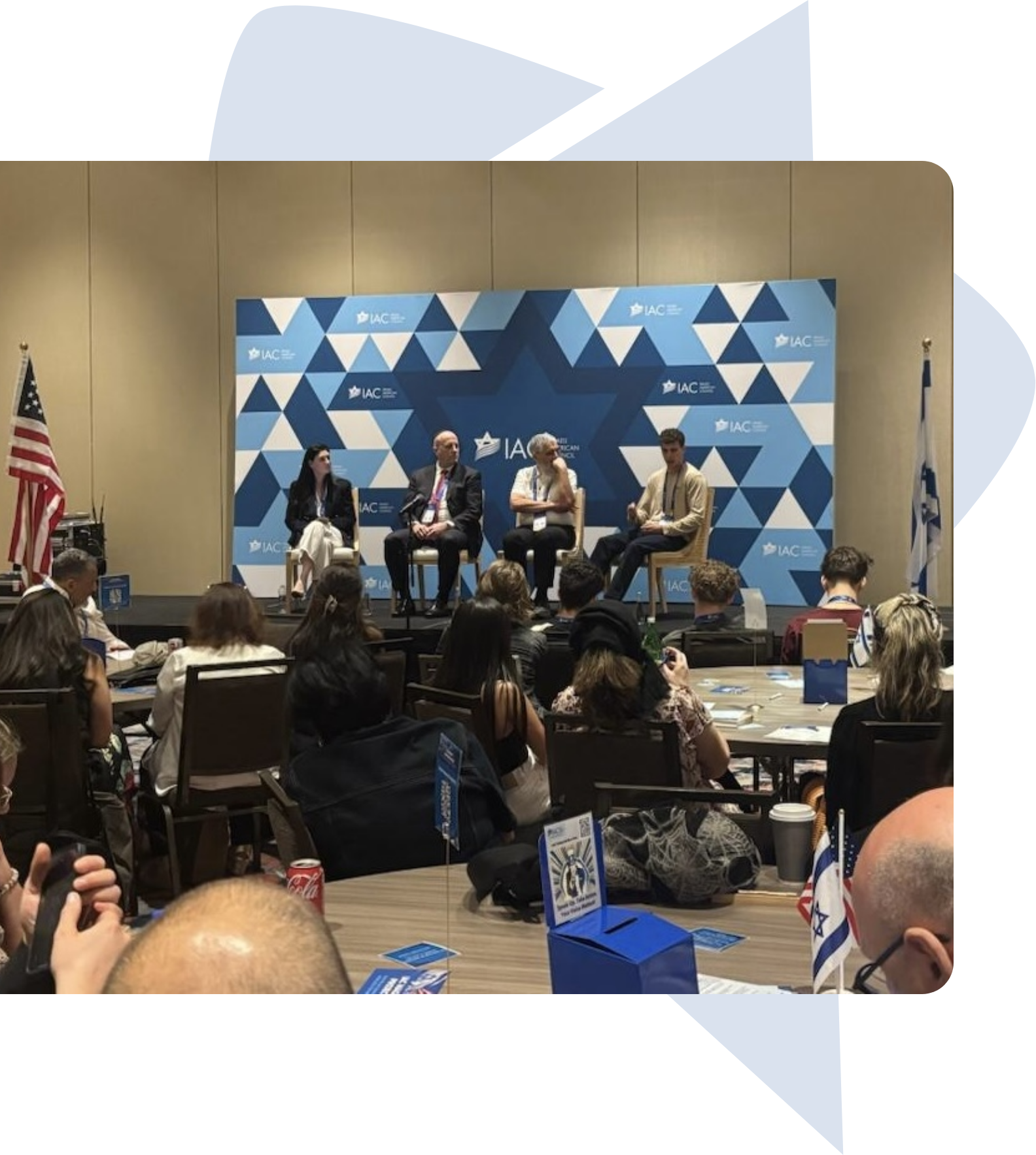 Panel discussion with four speakers seated on a stage with an audience in front, American and Israeli flags on either side, at an IAC event with a blue geometric star-themed backdrop.