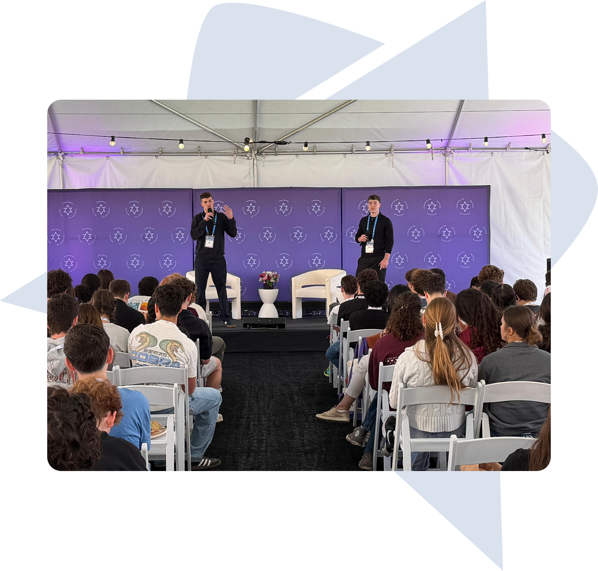 Two men standing on stage speaking to an audience seated in white chairs under a tent.