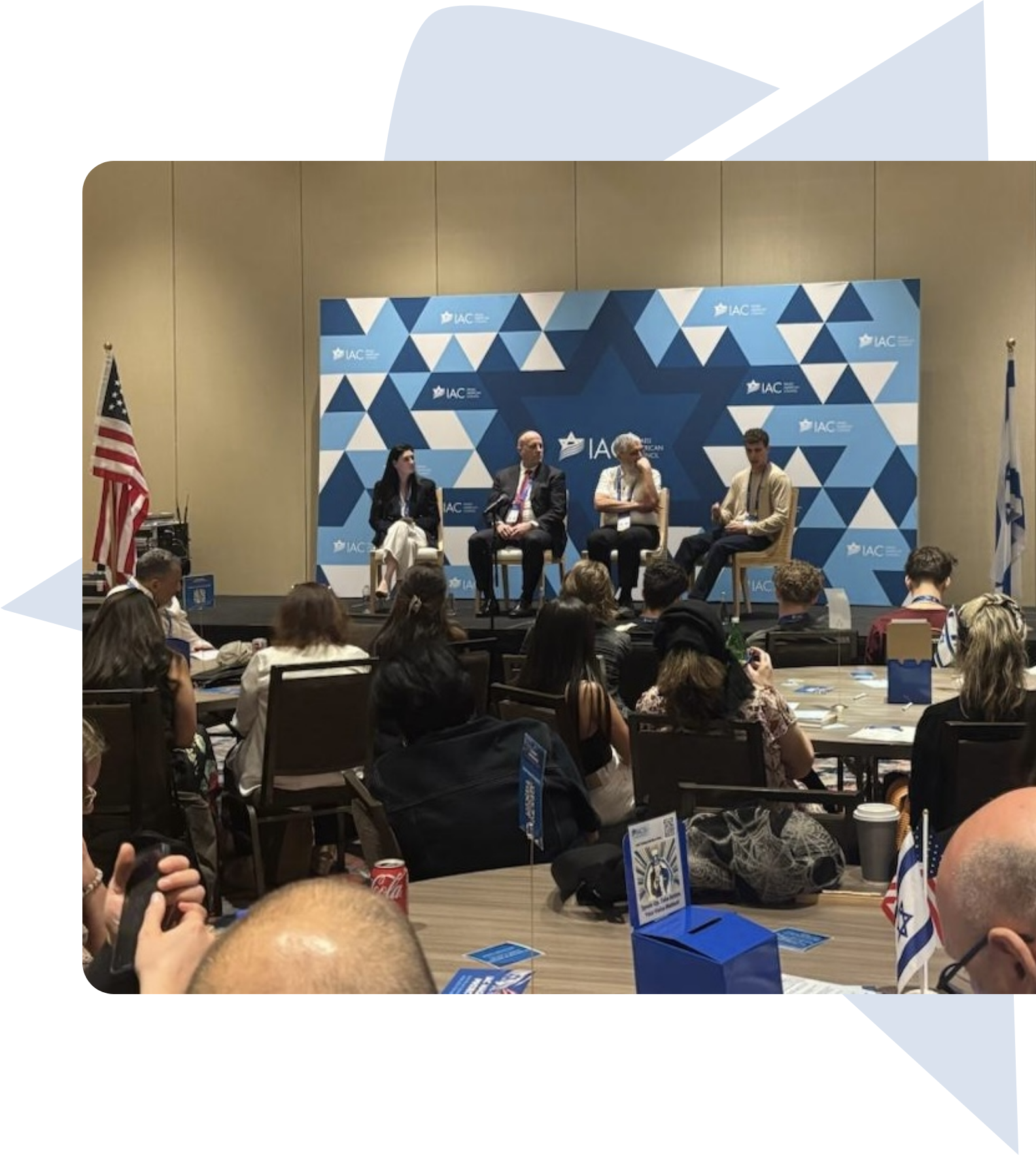 Panel discussion with four speakers seated on a stage with an audience in front, American and Israeli flags on either side, at an IAC event with a blue geometric star-themed backdrop.