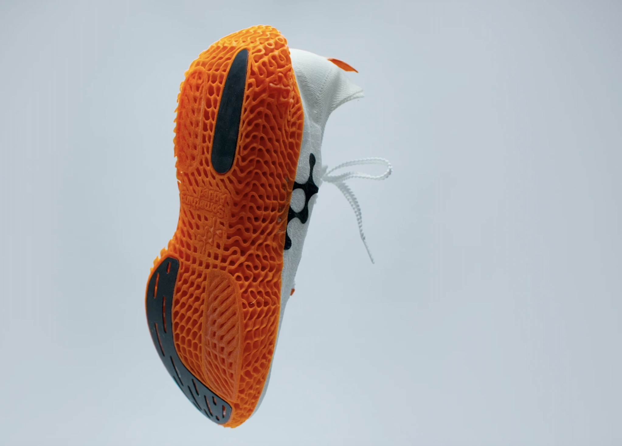 Close-up product shot of a white athletic sneaker by Something Added, suspended against a light gray background and shown from the sole view. The shoe features a bright orange, lattice-style outsole with black rubber grip accents and loose white laces hanging from the side. The modern sole design highlights Something Added’s distinctive textured construction and lightweight aesthetic.