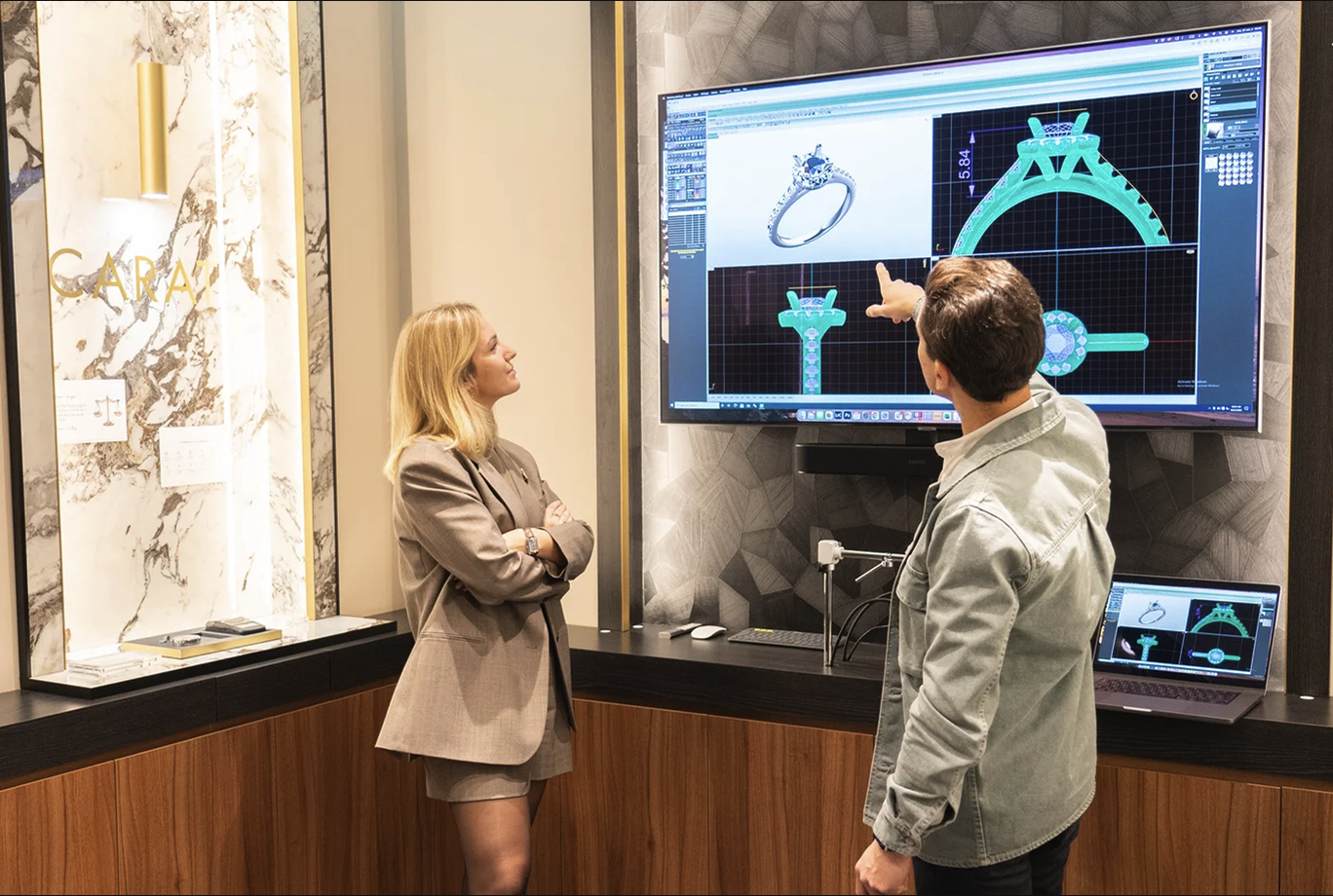 Two people from Amantys collaborate in a jewelry design showroom, standing in front of a large screen displaying detailed 3D renderings of a custom engagement ring. One person points to the digital design while the other watches attentively, reflecting Amantys’ collaborative approach to jewelry creation.