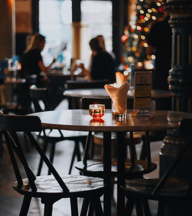 Runder Holztisch mit einem kleinen Kerzenhalter aus Glas und Servietten in einem Glas, in einem schwach beleuchteten Café mit verschwommenen Menschen und Weihnachtsbeleuchtung im Hintergrund.