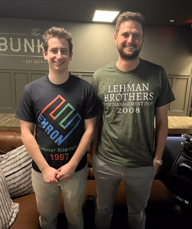Two men standing and smiling indoors, one wearing a black Enron Summer Internship 1997 t-shirt and the other wearing a green Lehman Brothers Risk Management Dept 2008 t-shirt.
