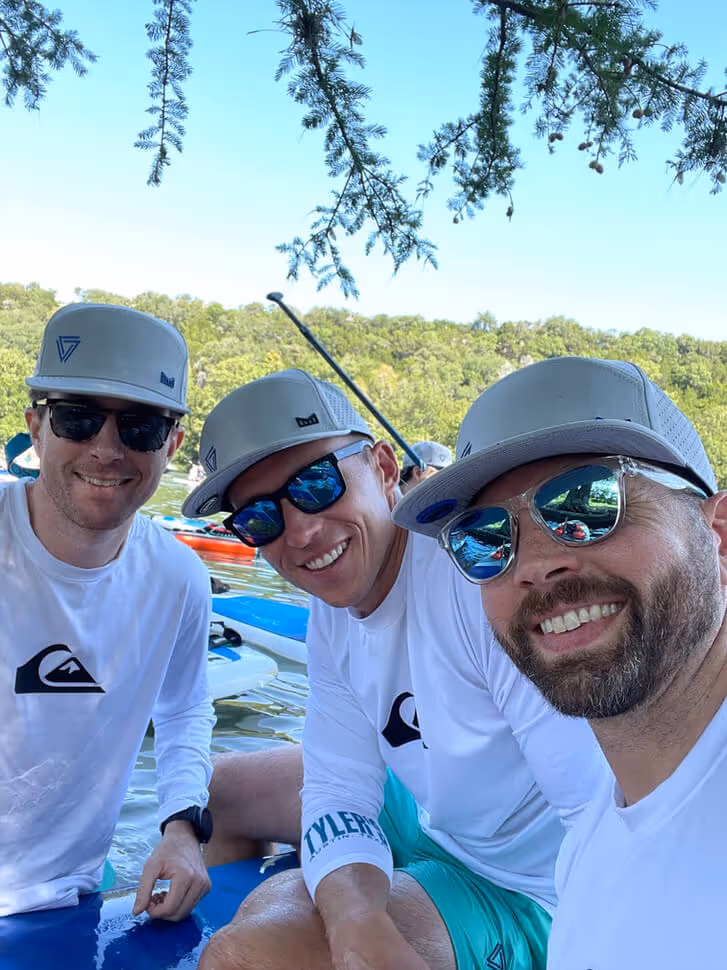 Three men wearing white long-sleeve shirts, teal shorts, gray caps, and sunglasses sitting on paddleboards on a lake surrounded by trees.