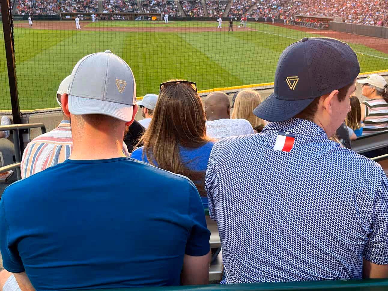 Two men wearing baseball caps seated in a stadium with a baseball game in progress on a green field in the background.