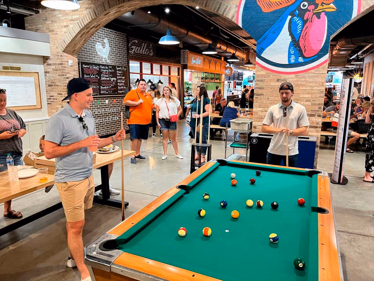 Two men playing pool at a billiards table inside a lively indoor market with people and food stalls in the background.
