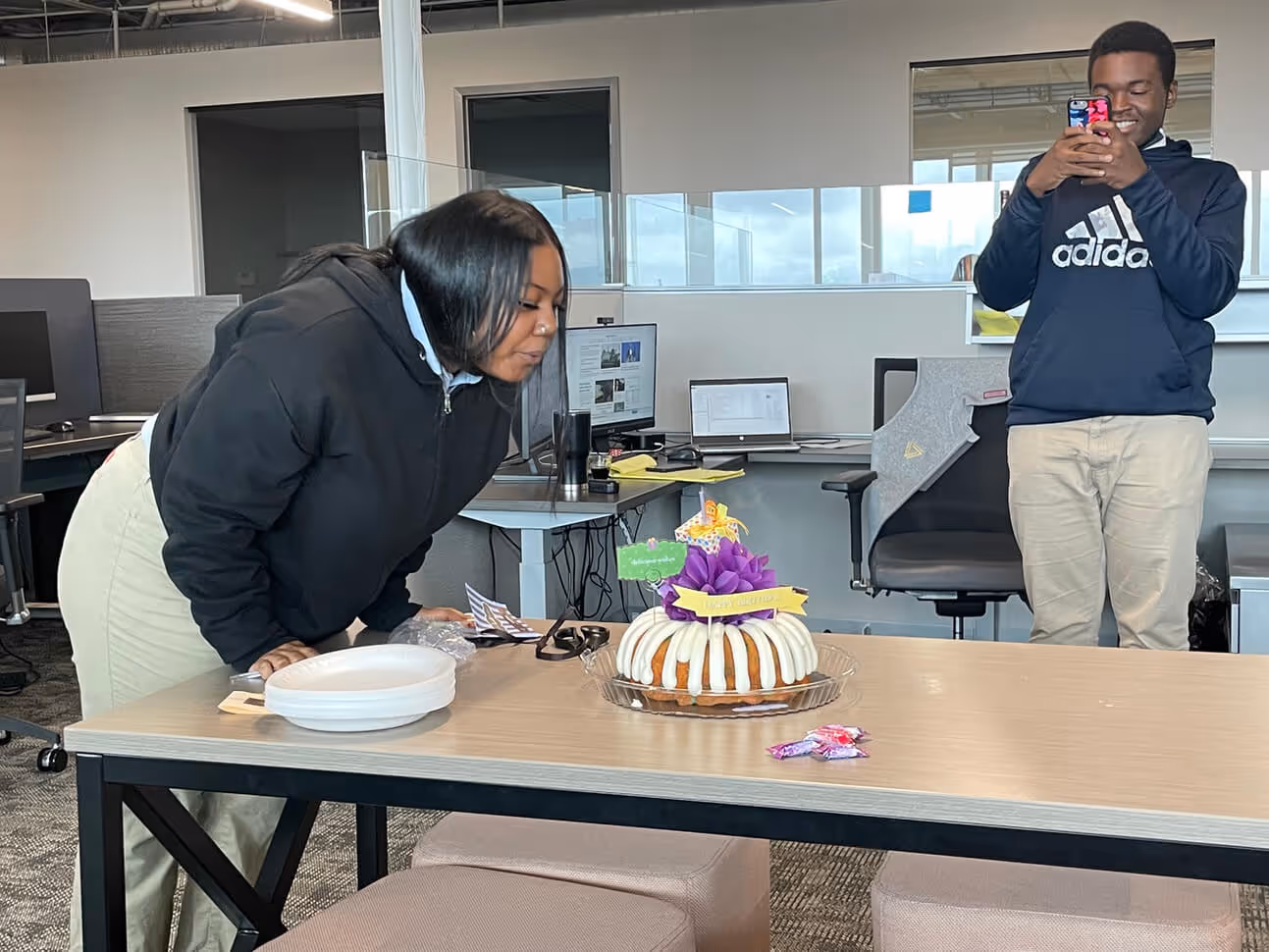 Woman leaning over a table blowing out a candle on a bundt cake with a man in an Adidas hoodie smiling and taking a photo in an office setting.