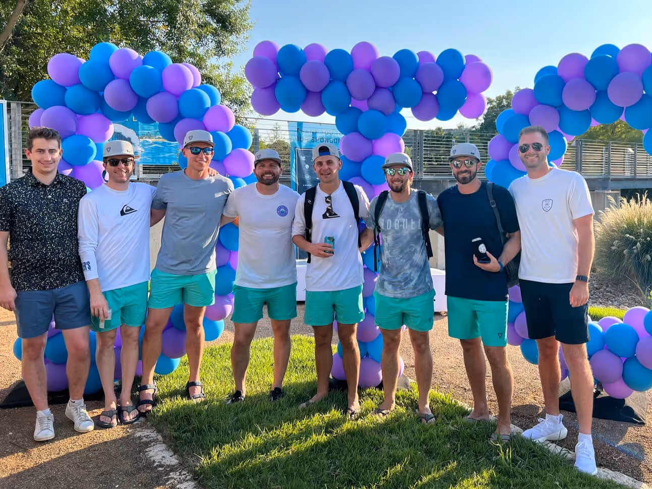Group of eight men standing outdoors in front of blue and purple balloon decorations, wearing casual summer outfits with several in matching teal shorts and hats.