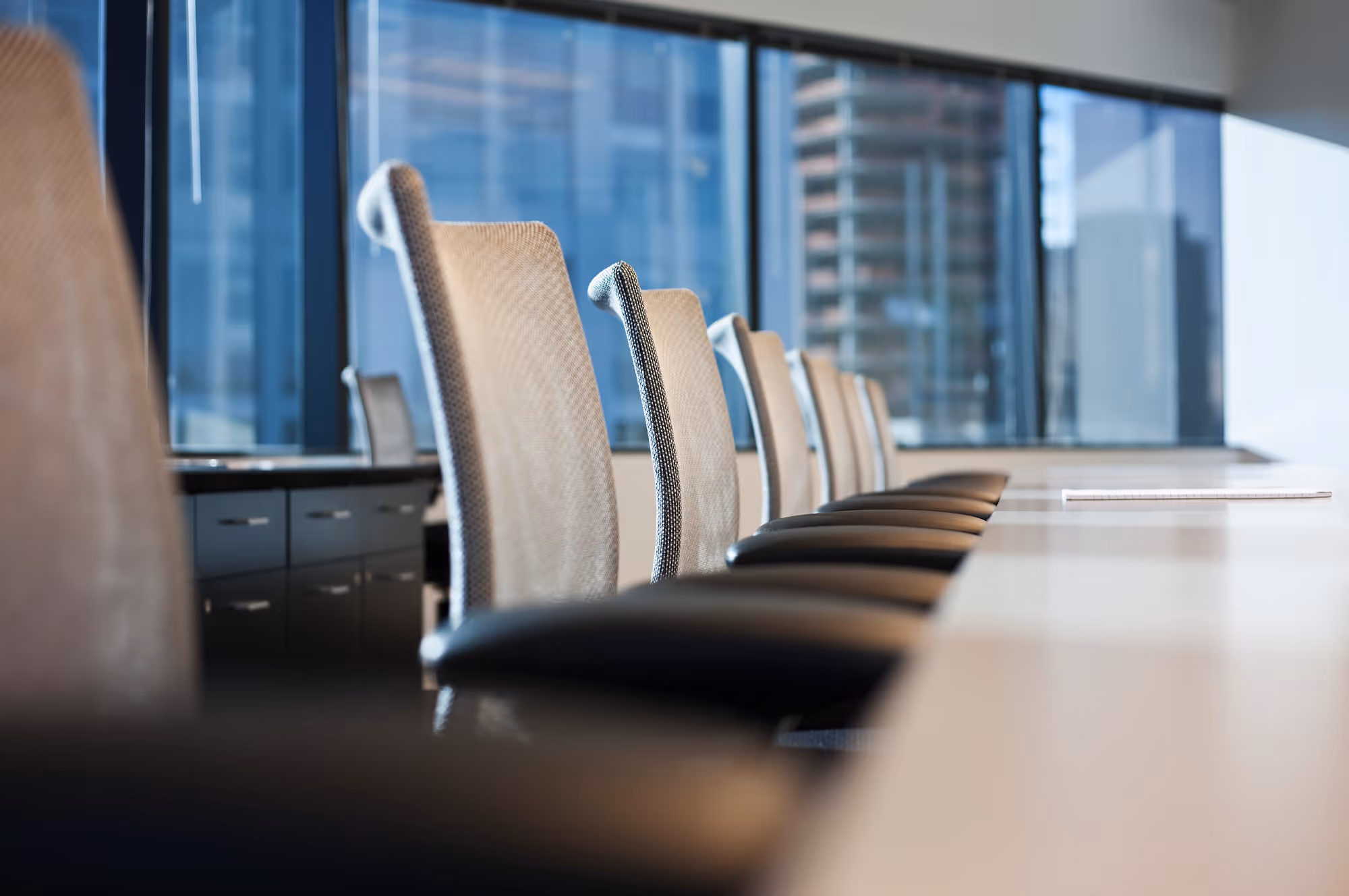 Row of empty mesh office chairs along a modern conference table with large windows overlooking a cityscape.
