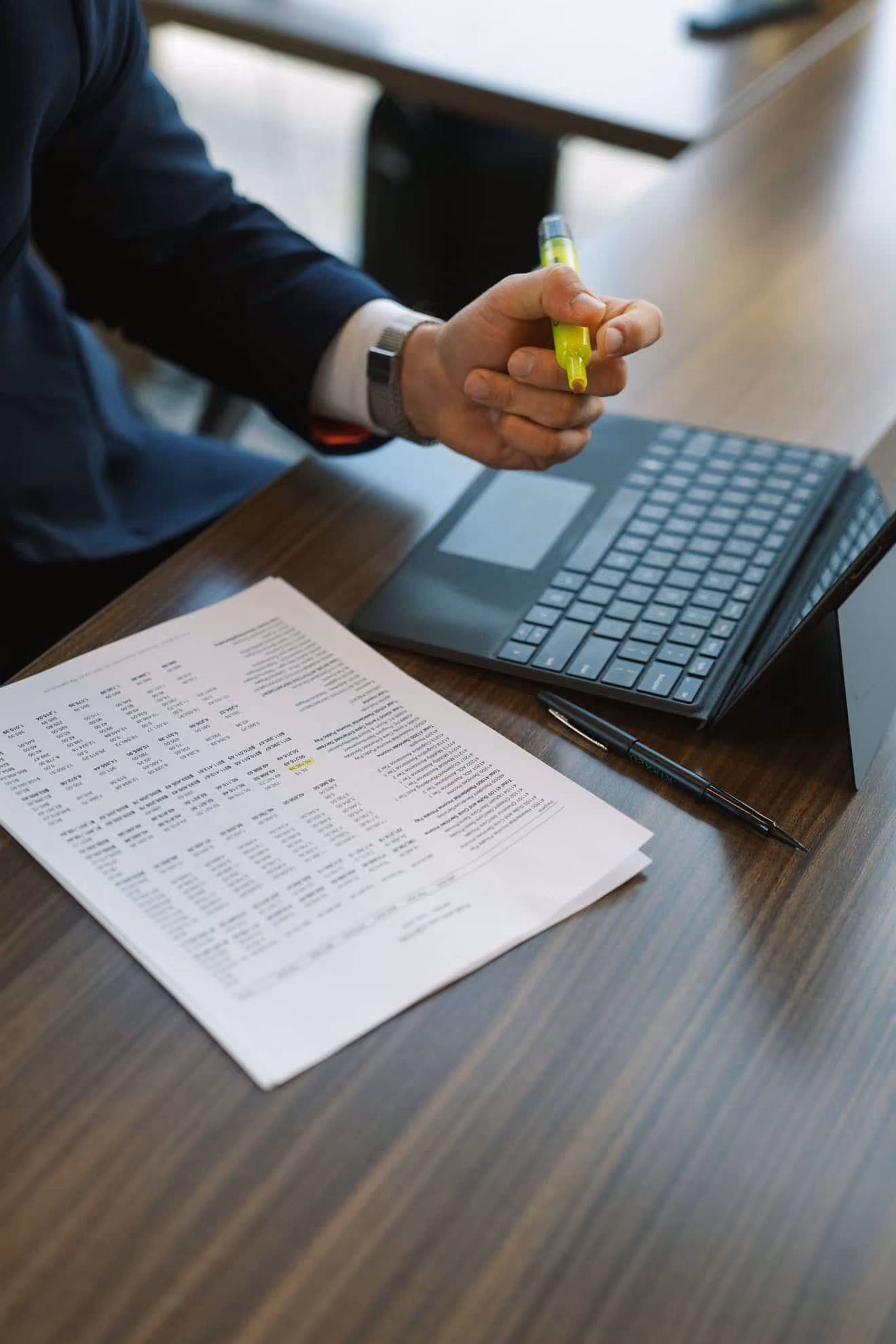 Person holding a yellow highlighter near a laptop on a wooden desk with printed documents and a black pen beside the keyboard.