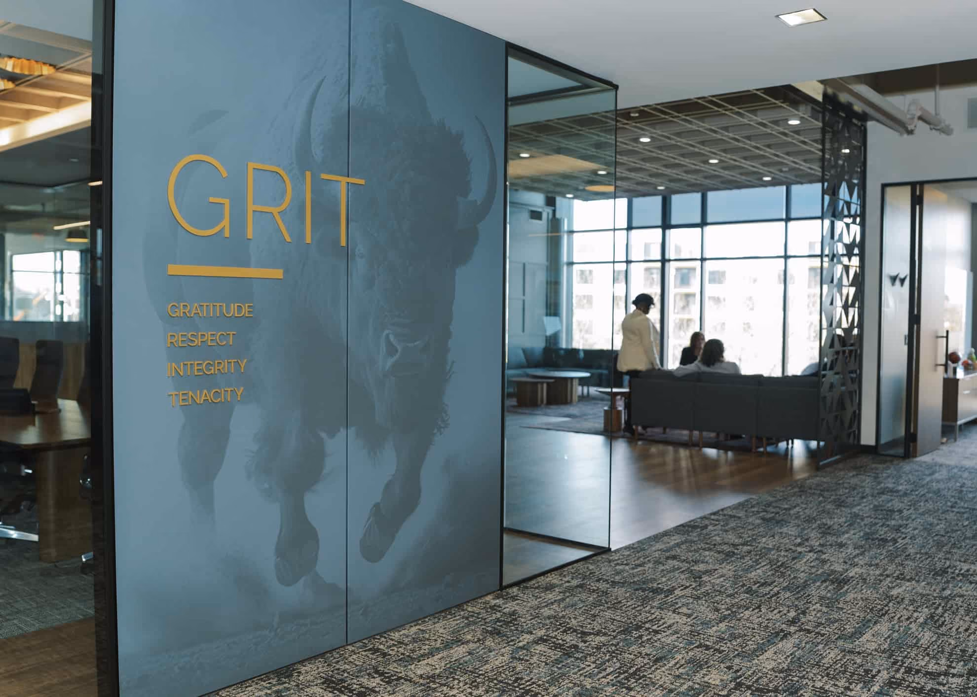 Office hallway with large glass wall featuring a buffalo image and the word GRIT with values: Gratitude, Respect, Integrity, Tenacity in yellow letters; people are sitting and standing in a glass-walled lounge area.