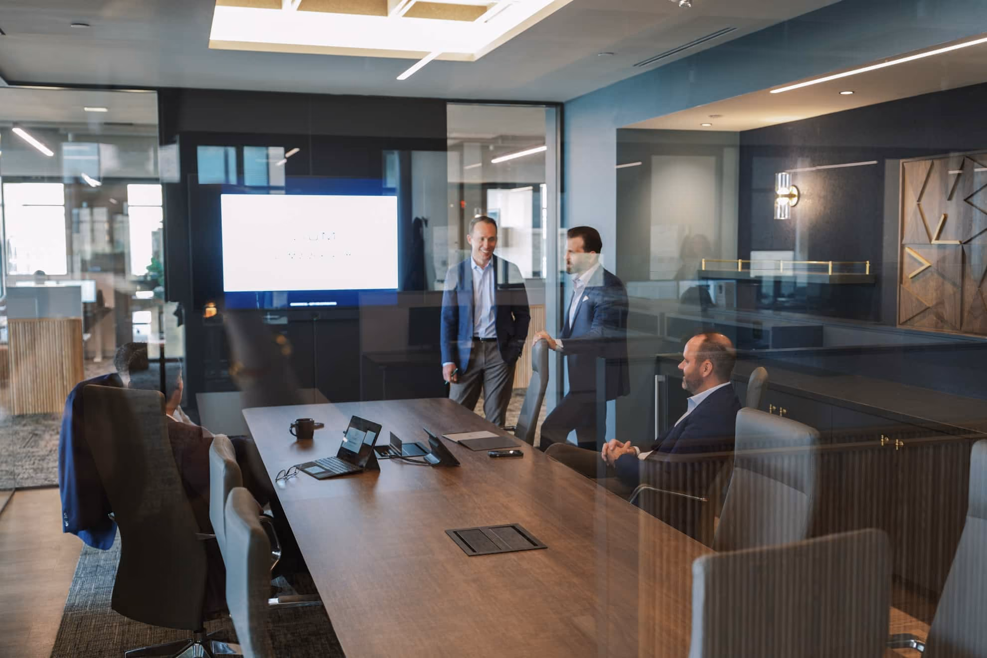 Four businessmen in a modern glass-walled conference room with laptops and calls devices on a large wooden table, two standing and two seated, engaged in discussion and smiling.