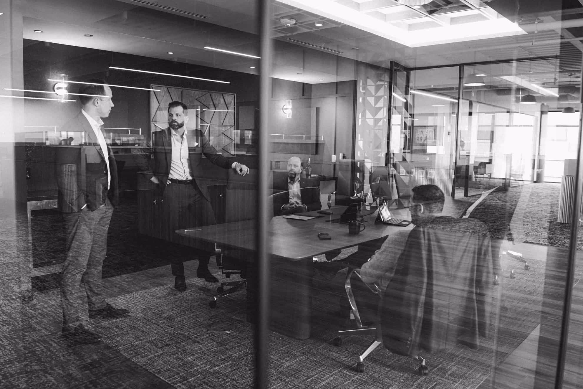 Black and white photo of four men in business attire having a meeting in a glass-walled conference room with a table, laptops, and office chairs.