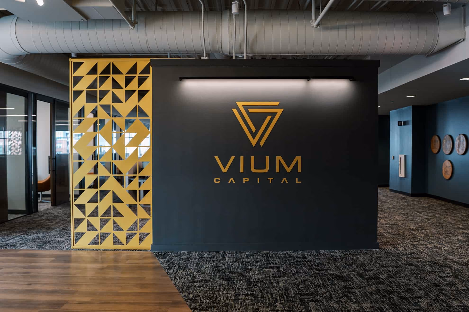 Modern office entry with a dark wall featuring the gold VIUM Capital logo, a geometric gold decorative panel to the left, and visible ductwork overhead.
