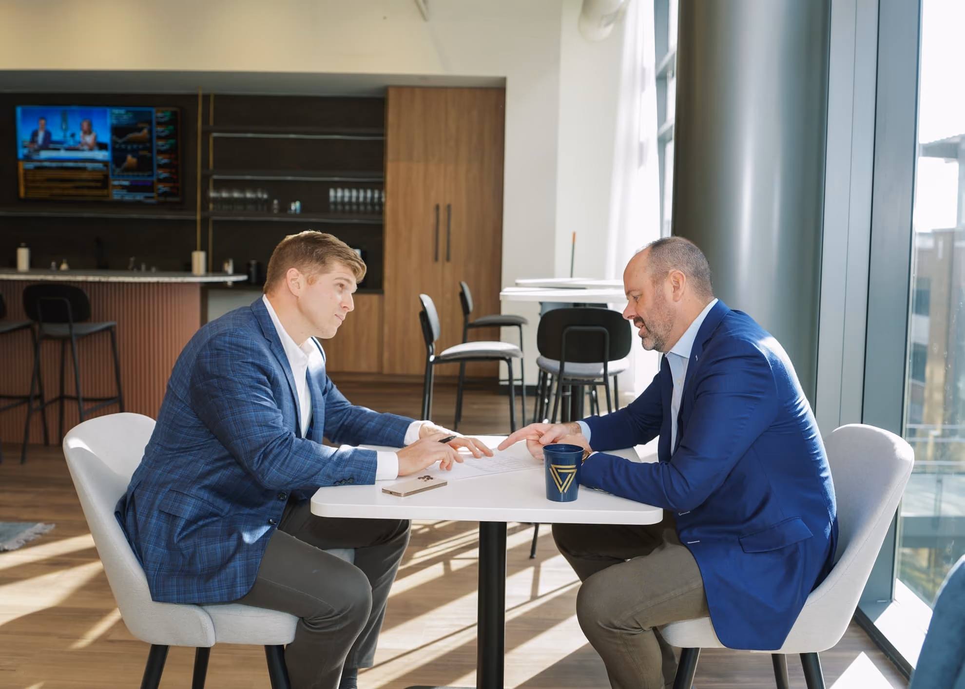 Two men in blue blazers sitting across a white table, discussing paperwork in a modern office with large windows and bar stools in the background.
