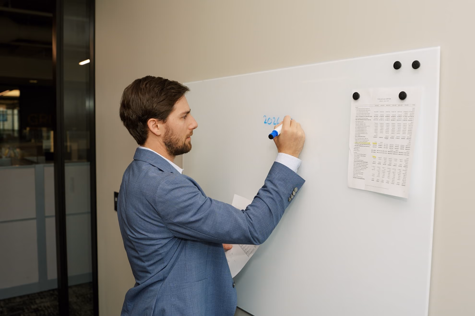 Man in a blue suit jacket writing '2026' on a whiteboard in an office, holding papers with printed tables pinned beside him.