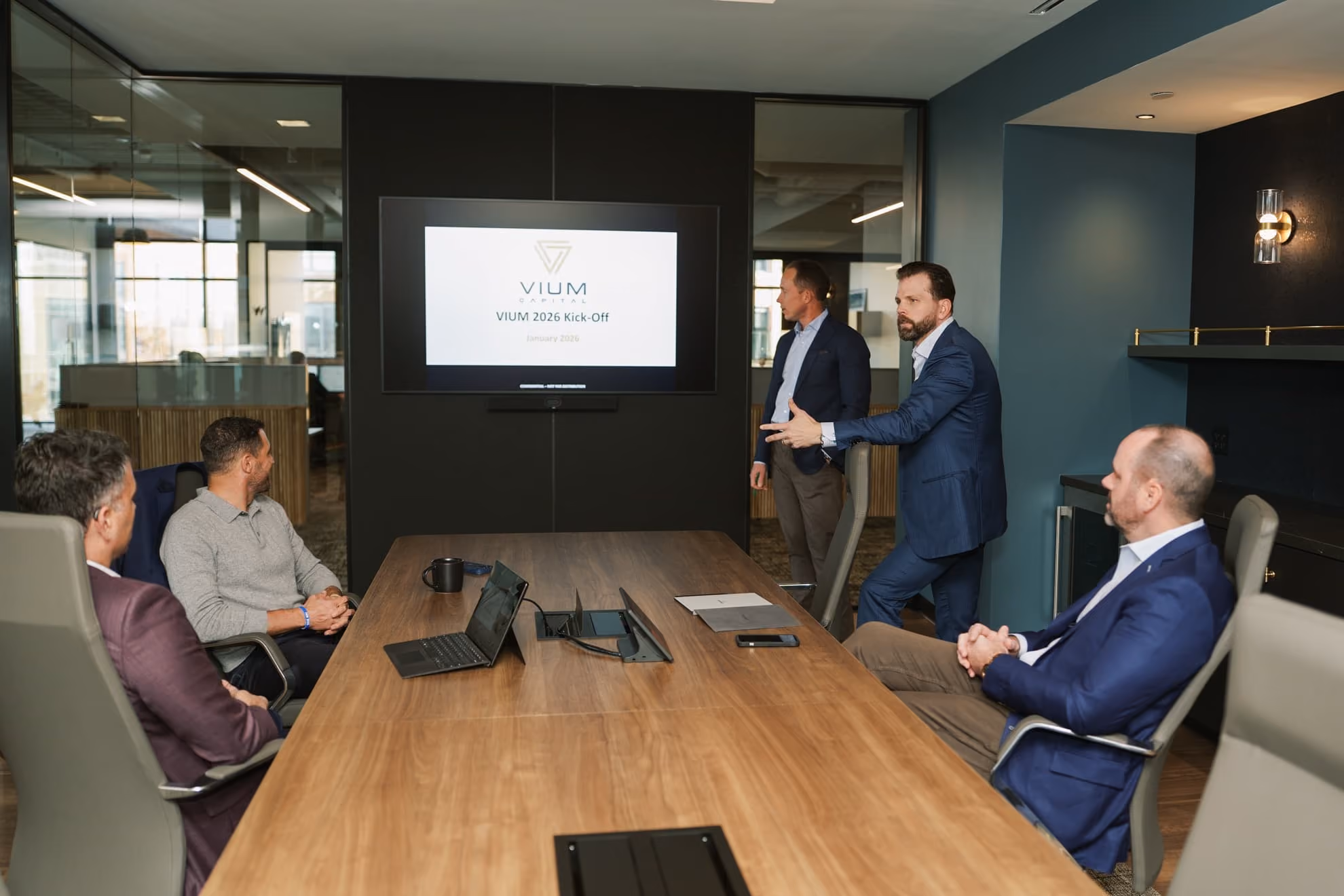 Five men in business attire in a conference room with one standing and pointing towards a screen displaying a presentation titled 'VIUM 2026 Kick-Off'.
