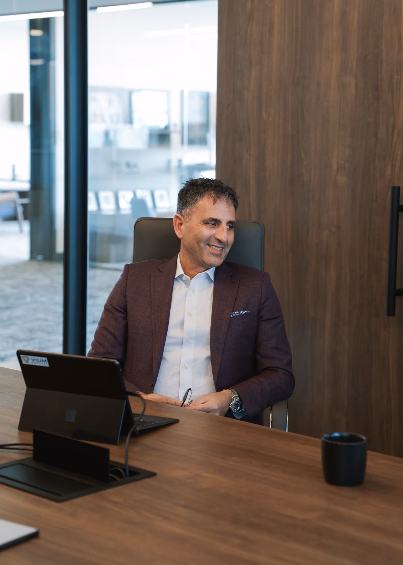 Man in a purple blazer and white shirt sitting at a wooden conference table, smiling and holding glasses, with a laptop and a black cup on the table in a modern office setting.