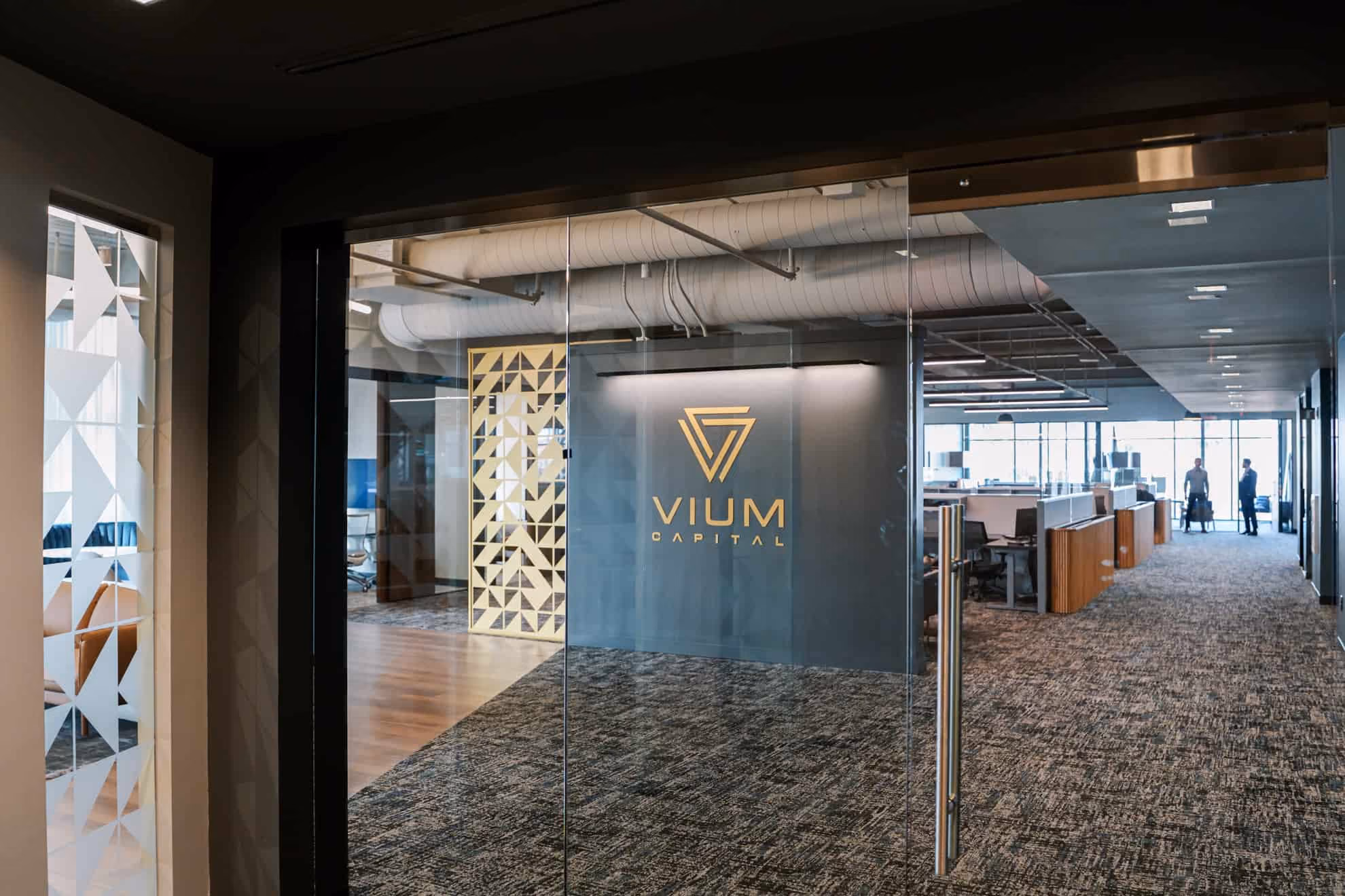 Modern office interior with glass entrance door featuring the Vium Capital logo, open workstations, a geometric patterned wall divider, and two people standing in the background near large windows.