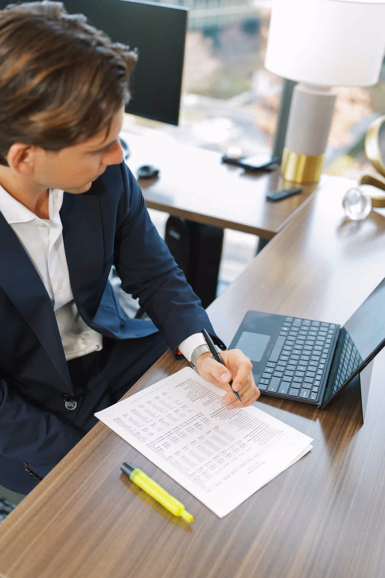Man in a navy suit sitting at a wooden desk, writing on a document with printed tables, next to an open laptop and a yellow highlighter.