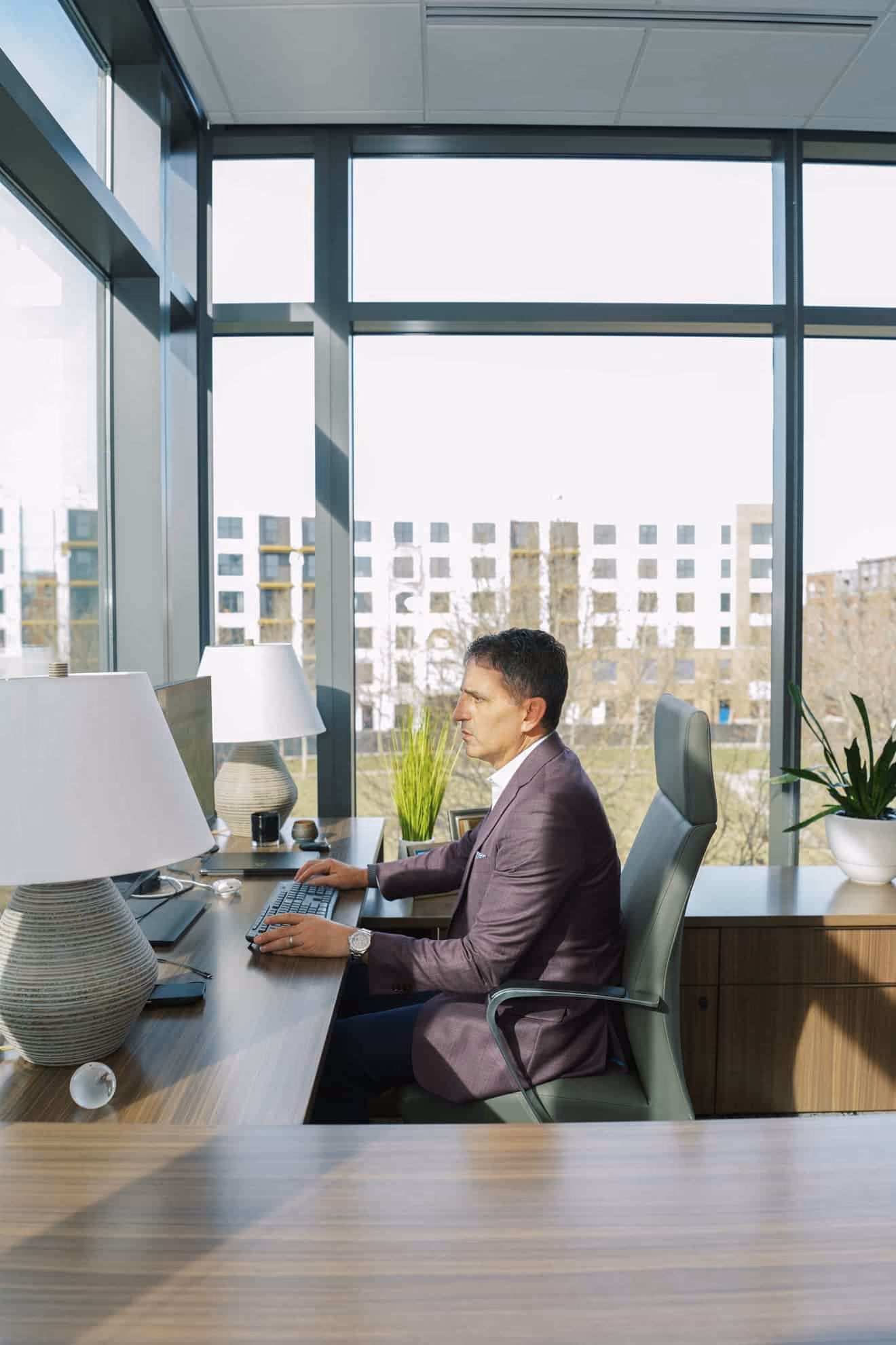 Man in a purple blazer working on a desktop computer in a modern office with large windows, plants, and a wooden desk.
