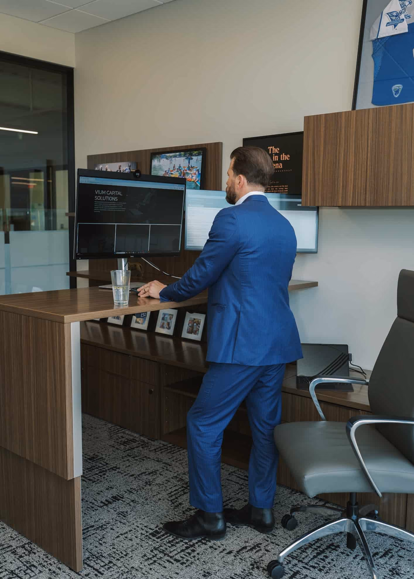 Man in a blue suit standing at a wooden desk in an office, working on a computer with dual monitors, a glass of water on the desk, and office decor including framed photos and a sports jersey on the wall.
