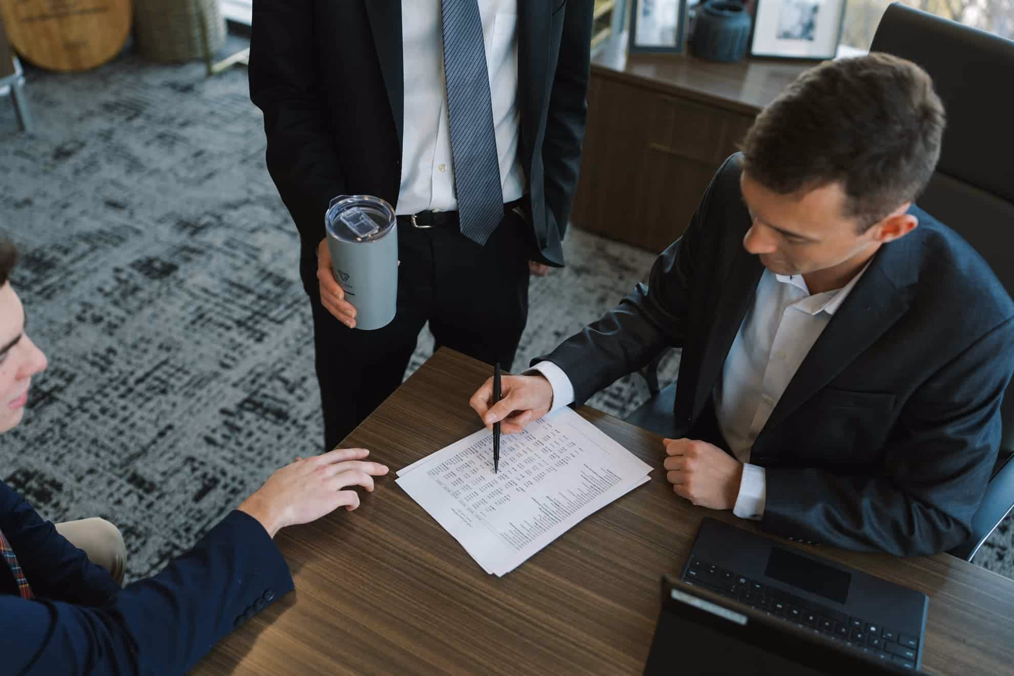 Three men in business attire at a table; one seated man points with a pen to a document while another seated man reaches toward it and a standing man holds a travel mug.