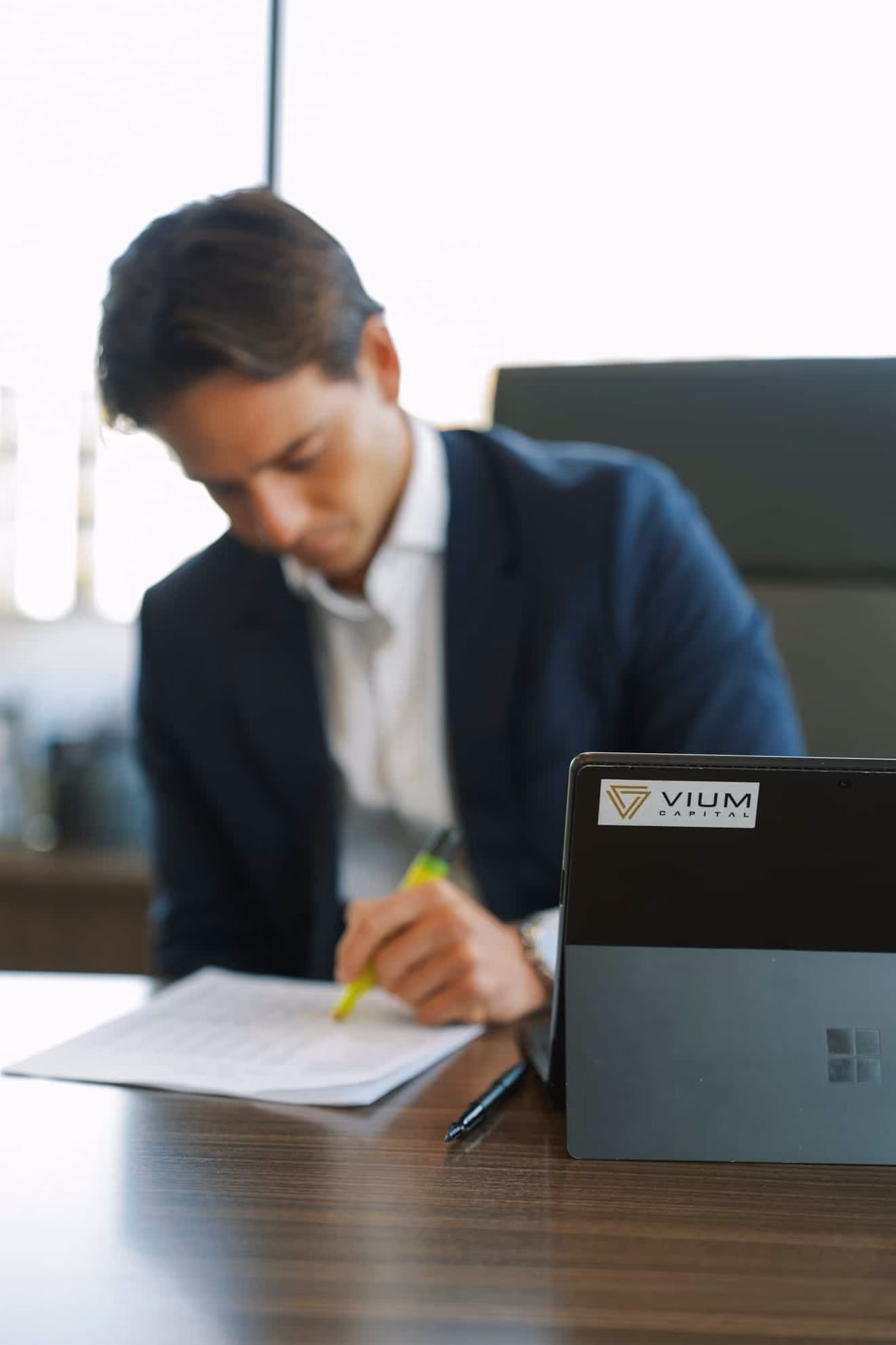 Businessman in a suit highlighting text on a document at a desk with a laptop in the foreground displaying a VIUM Capital logo.