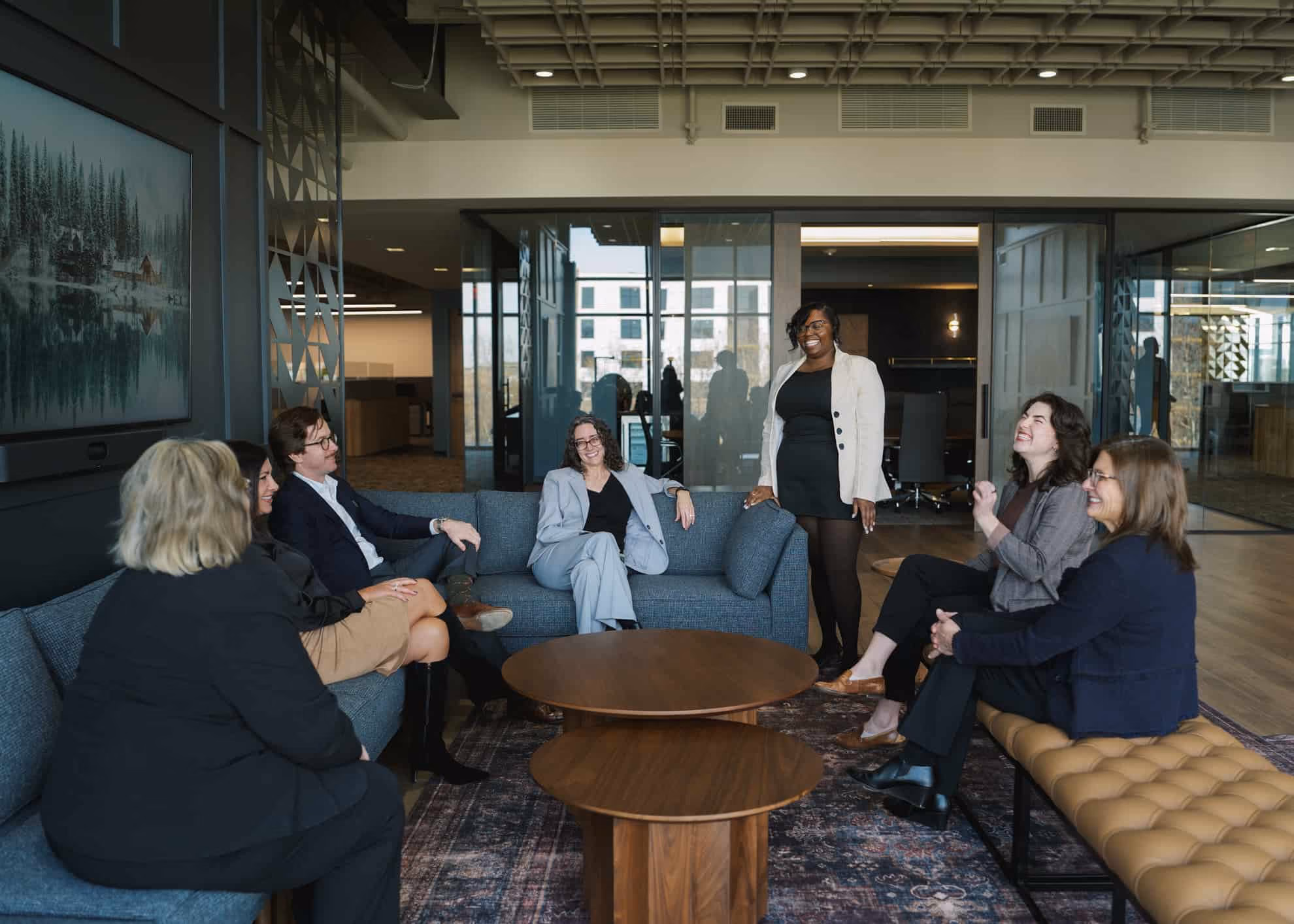 Seven professionals smiling and conversing in a modern office lounge with blue sofas, a tan bench, and two round wooden tables on a patterned rug.
