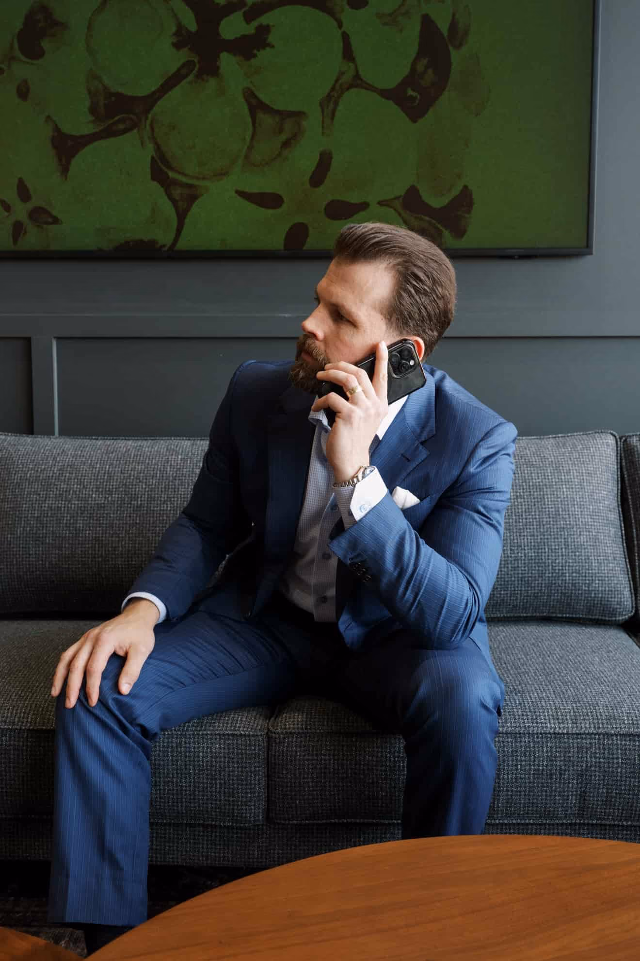 Man in a blue suit sitting on a gray couch, holding a phone to his ear with a thoughtful expression.
