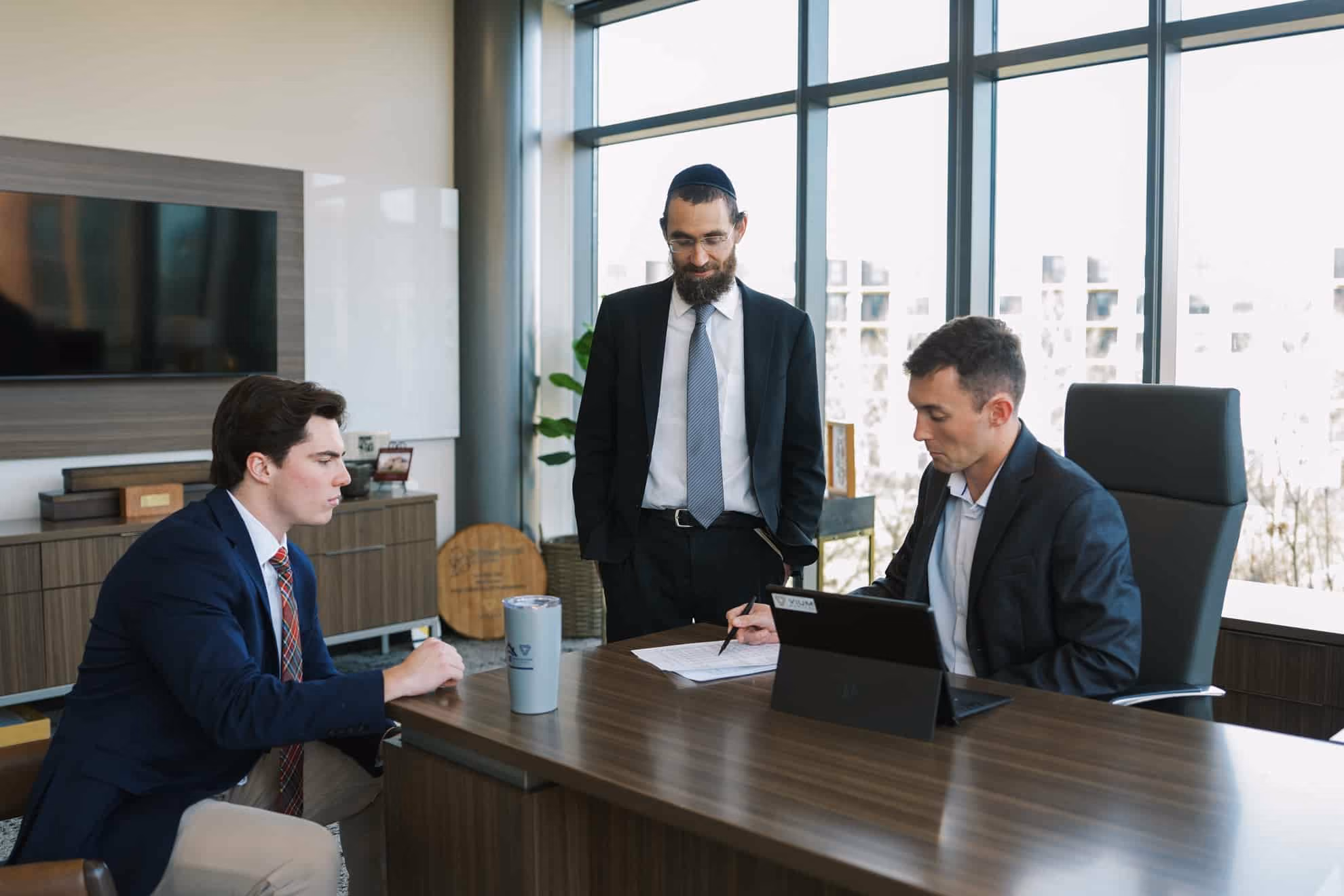 Three men in an office meeting: one seated in front of a laptop signing papers, one standing watching, and one seated listening with a drink on the table.
