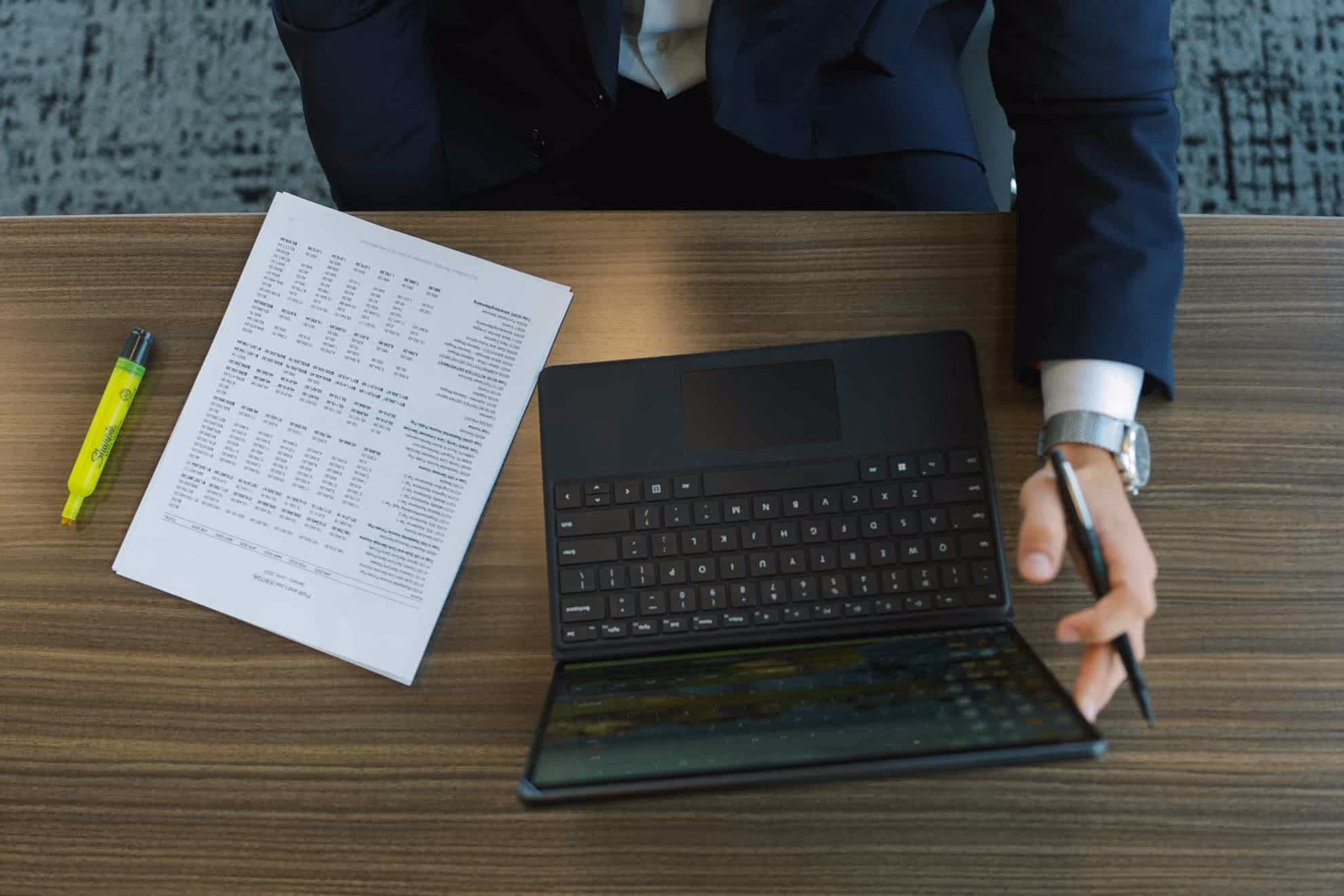 Person in a dark suit sitting at a wooden table with an open tablet keyboard, a pen in hand, a watch on the wrist, a yellow highlighter, and printed sheets with tables and text.