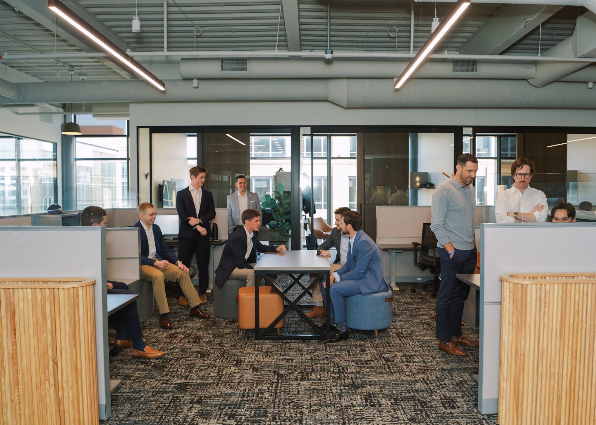 Group of men in business attire gathered and interacting in a modern office space with cubicles, a table, and large windows in the background.