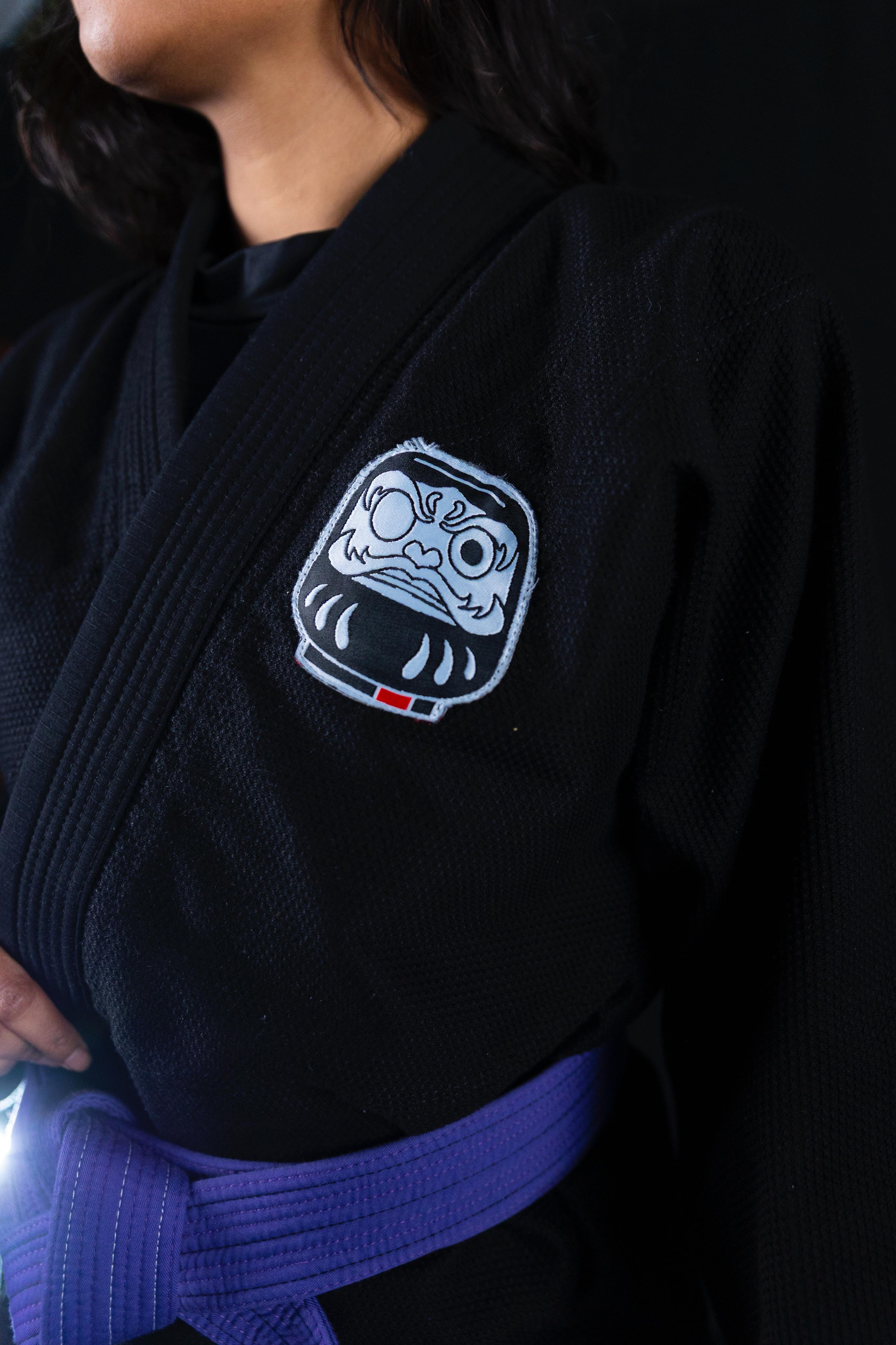 Person wearing a black martial arts gi with a purple belt and a white Daruma doll patch on the chest.