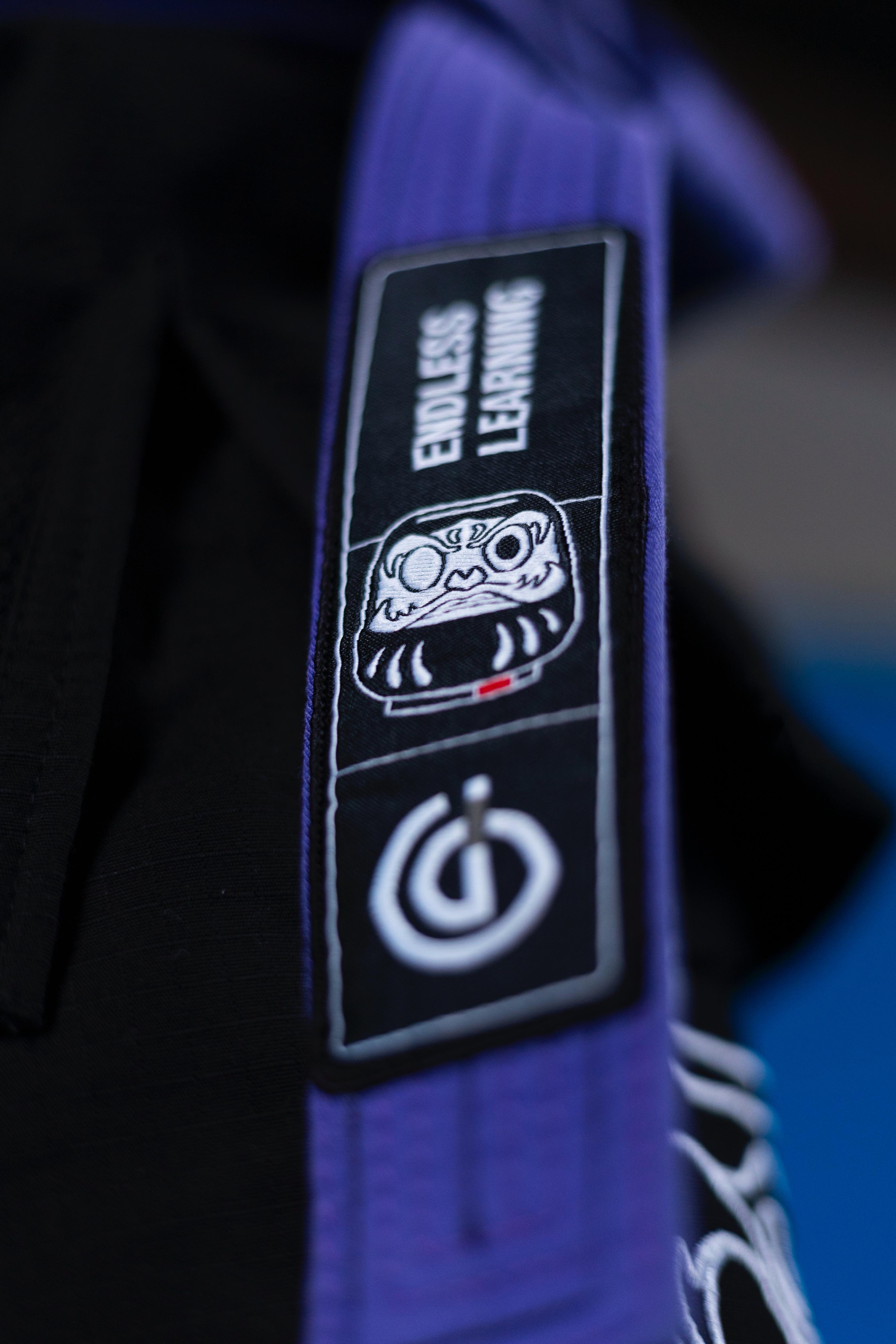 Close-up of a purple martial arts belt with a patch featuring a Daruma doll face, the text 'ENDLESS LEARNING,' and a power button symbol.