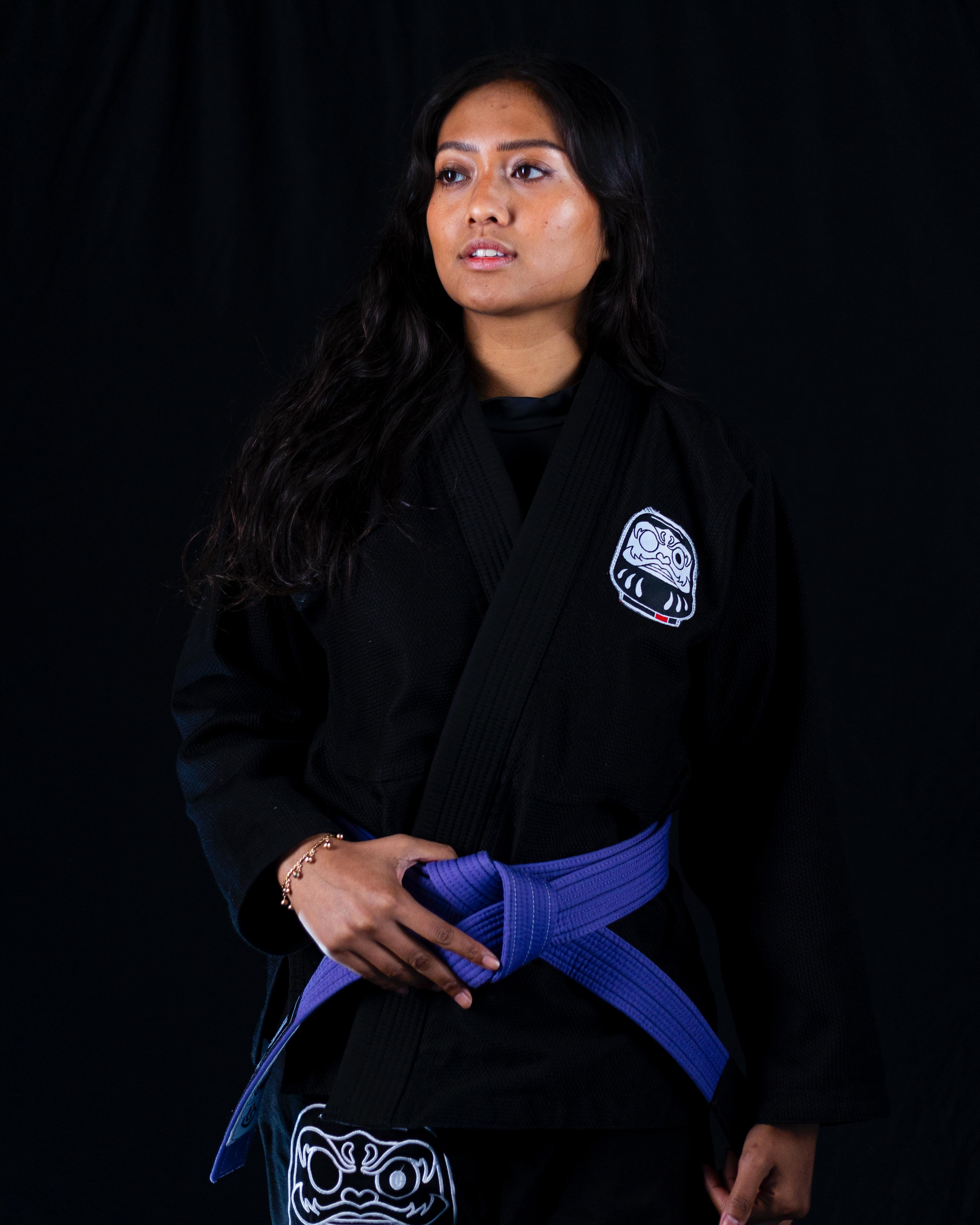 Woman wearing a black martial arts gi with a blue belt tied around her waist, standing against a black background.