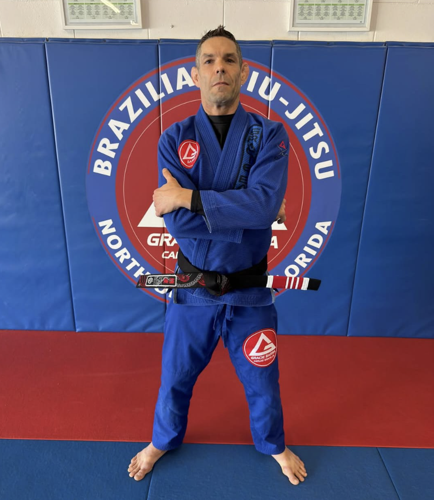 Man in a blue Brazilian Jiu-Jitsu gi with a black belt stands barefoot on blue and red mats with arms crossed in front of a Brazilian Jiu-Jitsu academy logo wall padding.