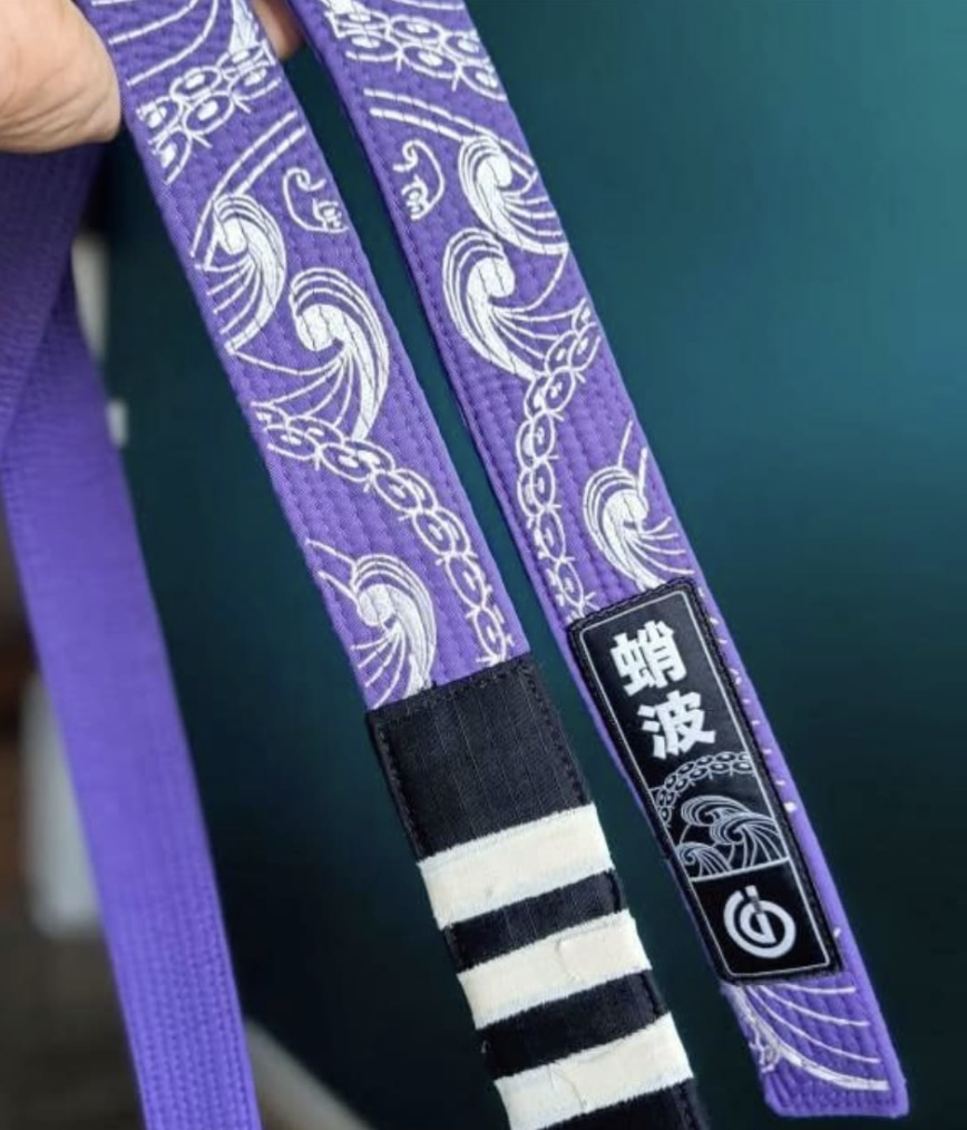 Close-up of a purple martial arts belt with white wave patterns and a black label with white Japanese characters and a logo.