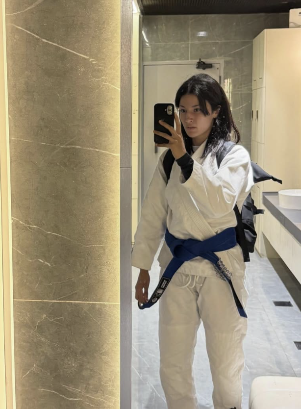 Young woman wearing a white martial arts gi with a blue belt taking a mirror selfie in a bathroom.